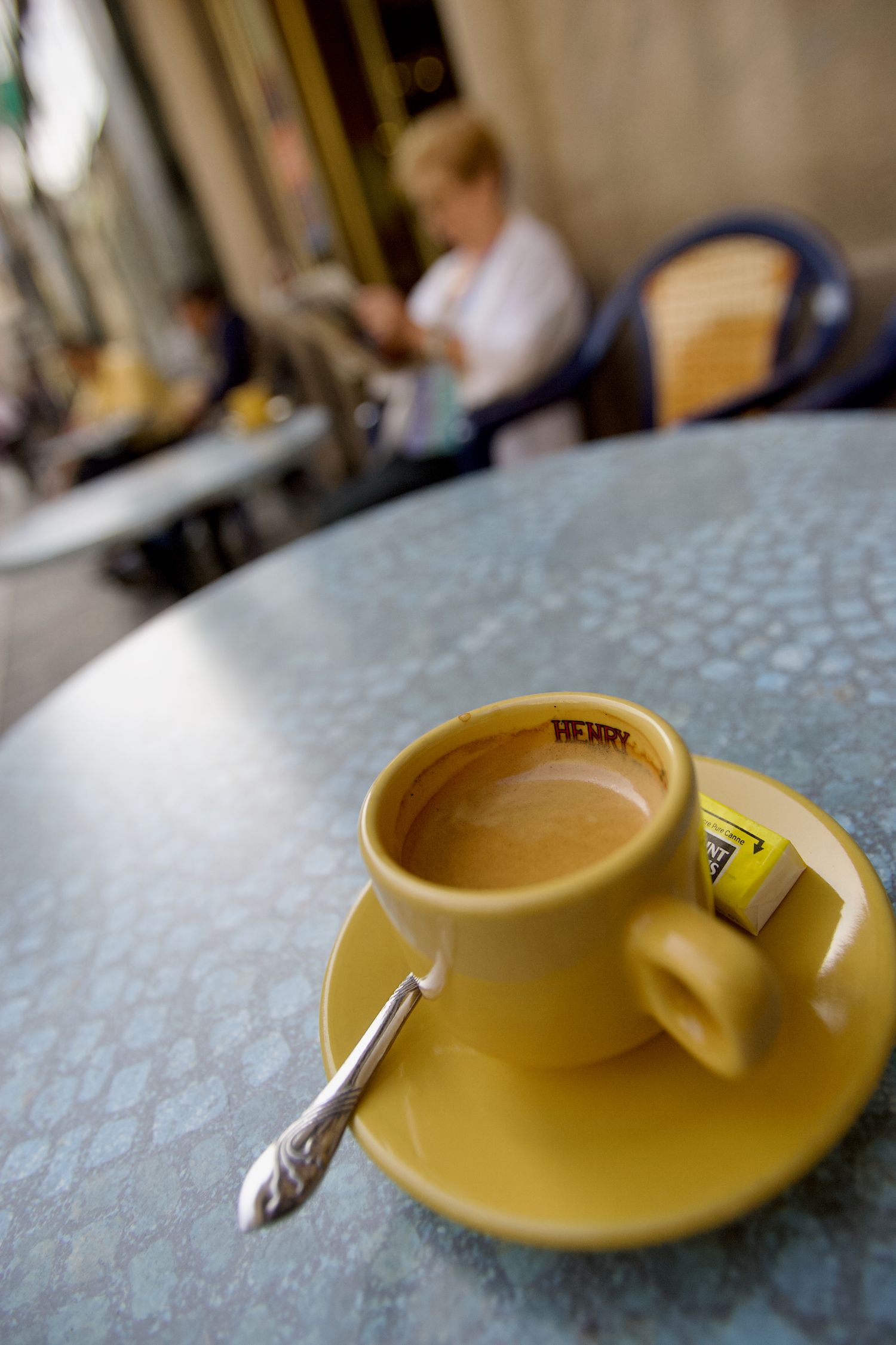 A yellow coffee cup and saucer sits on a textured blue tablecloth in a cafe setting.