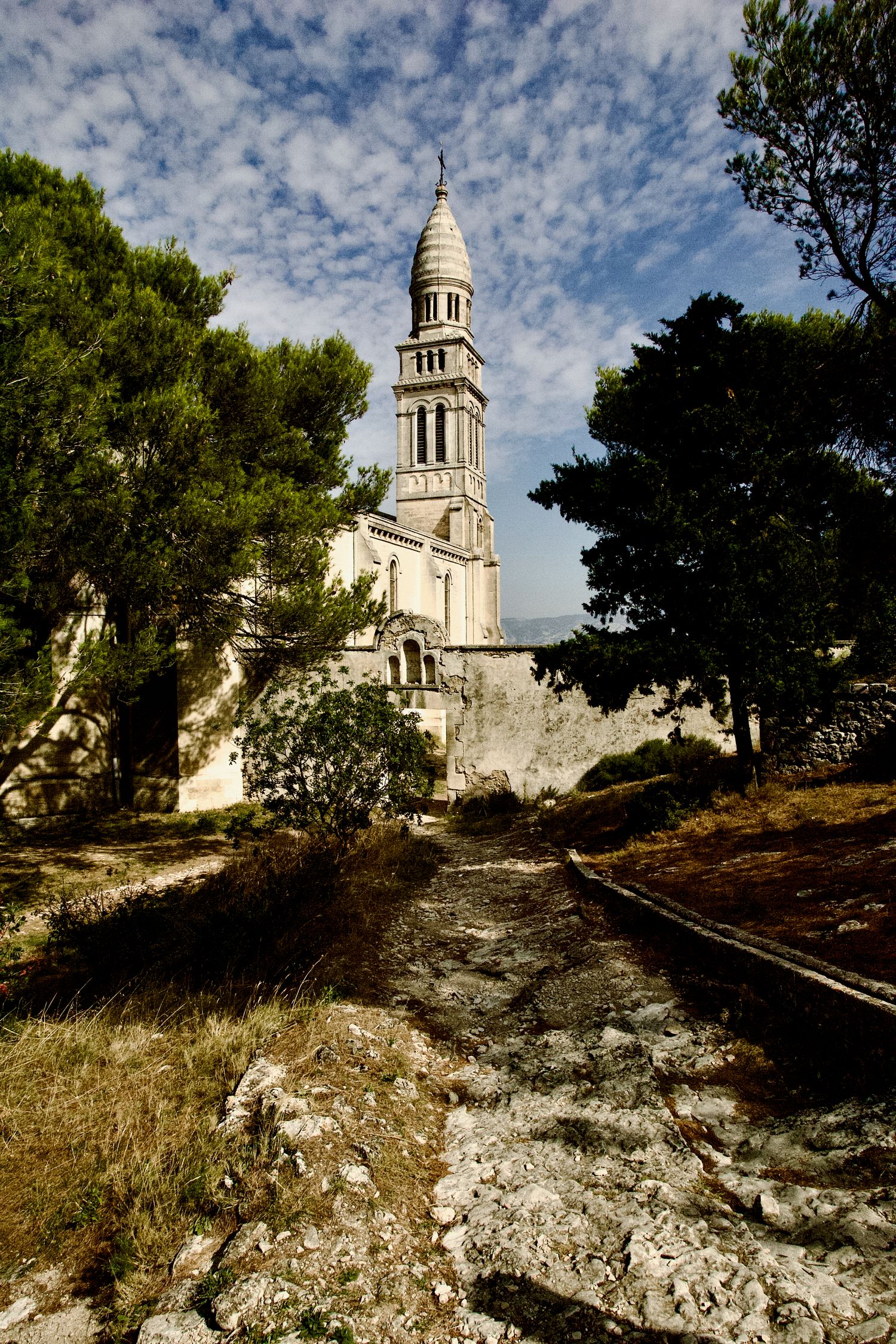 White stone church with tower stands majestically on rocky Mediterranean hillside under cloudy sky.