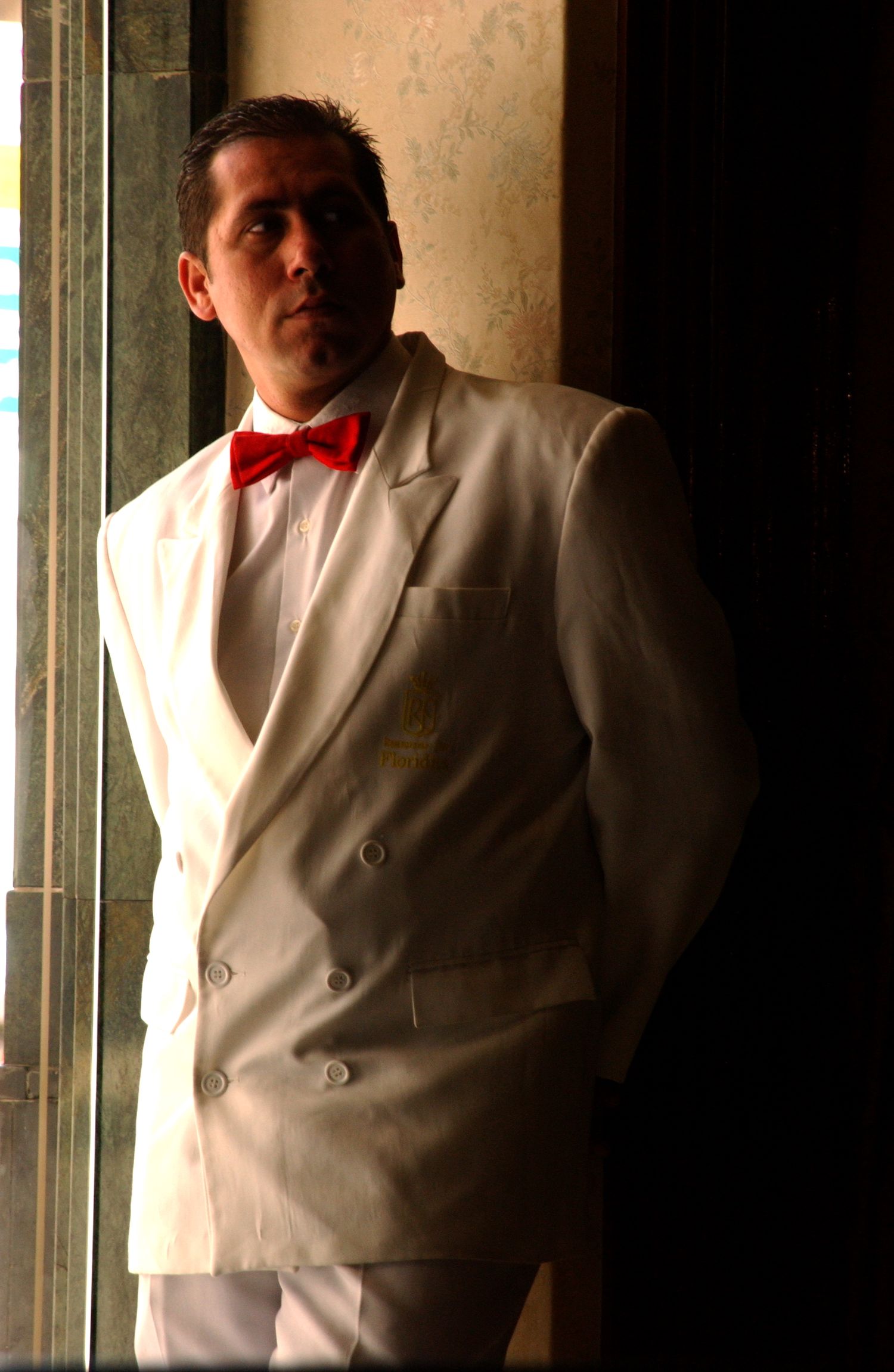 Person wearing a white suit jacket and red bowtie poses in dimly lit interior.