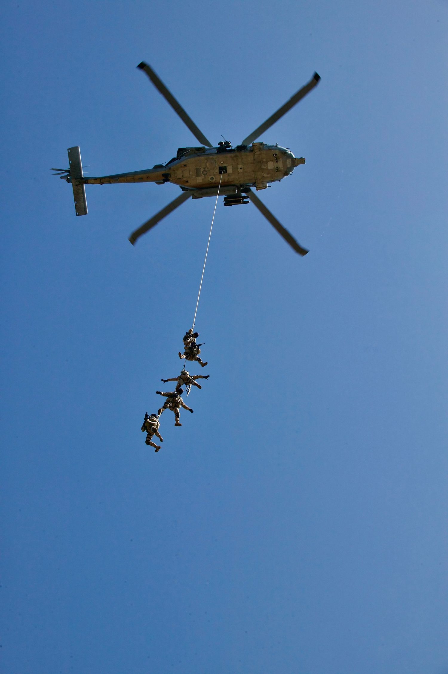 Military helicopter performs aerial training exercise with soldiers rappelling down ropes against clear blue sky.
