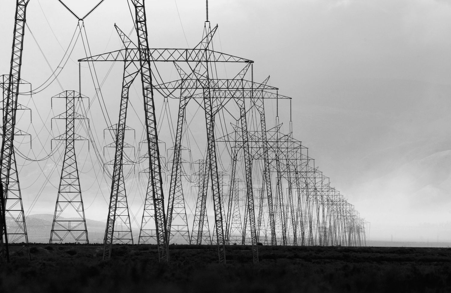Silhouettes of electrical transmission towers and power lines stretch across a moody sky in black and white.