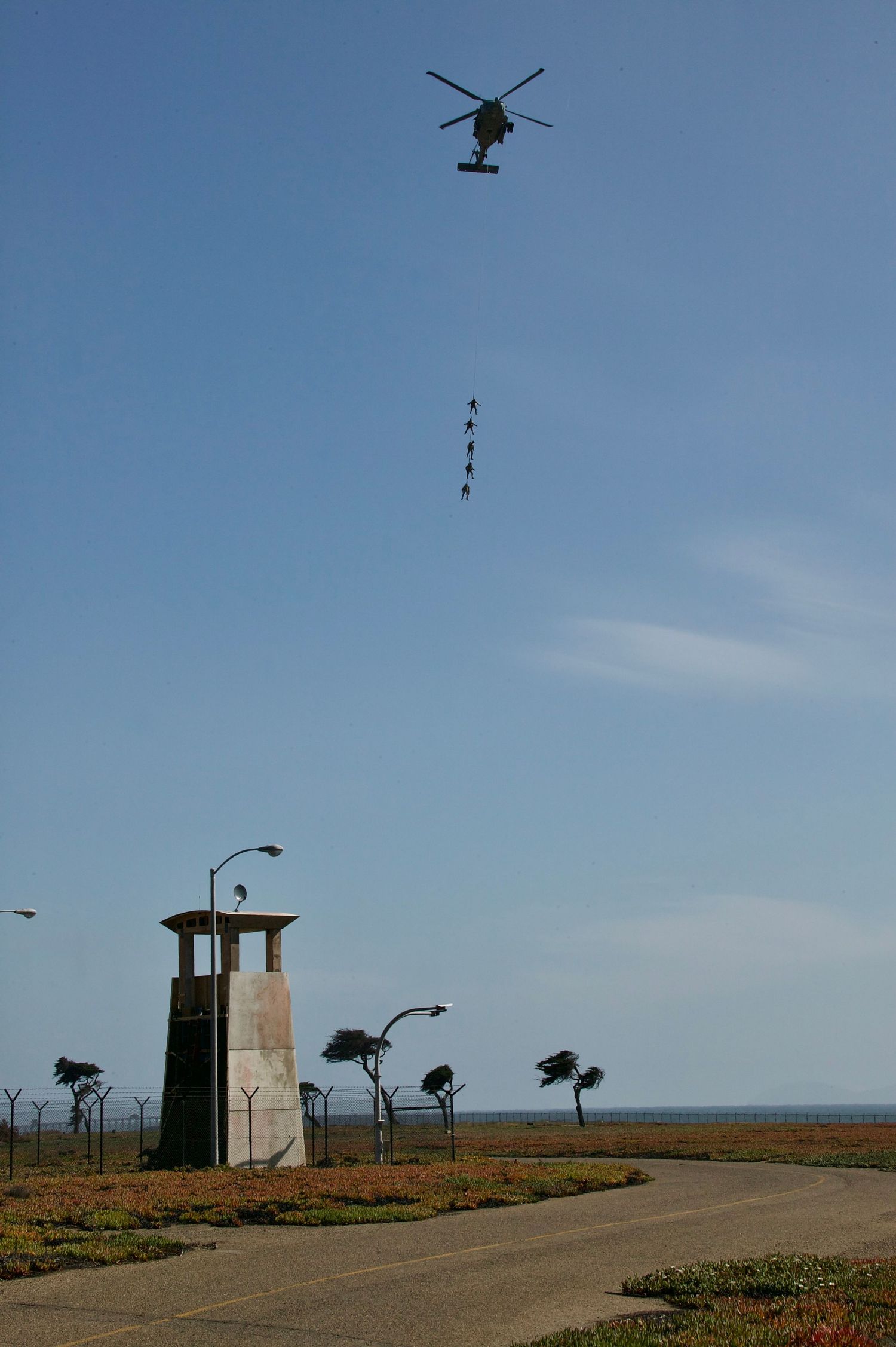 A helicopter flies over a coastal road with a lookout tower and palm trees against a blue sky.