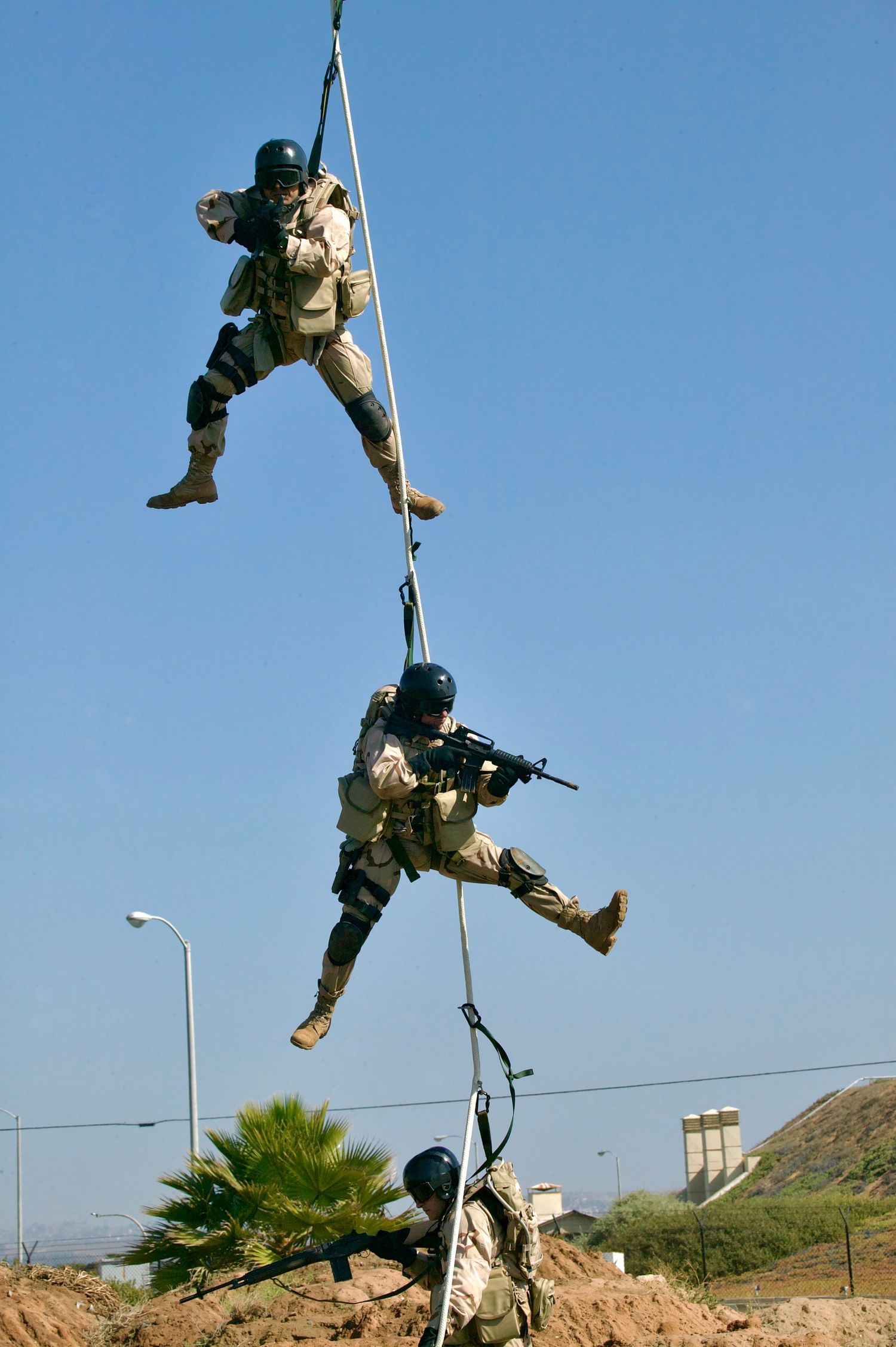 Military personnel rappel down a rope line during a training exercise against a bright blue sky and desert landscape.