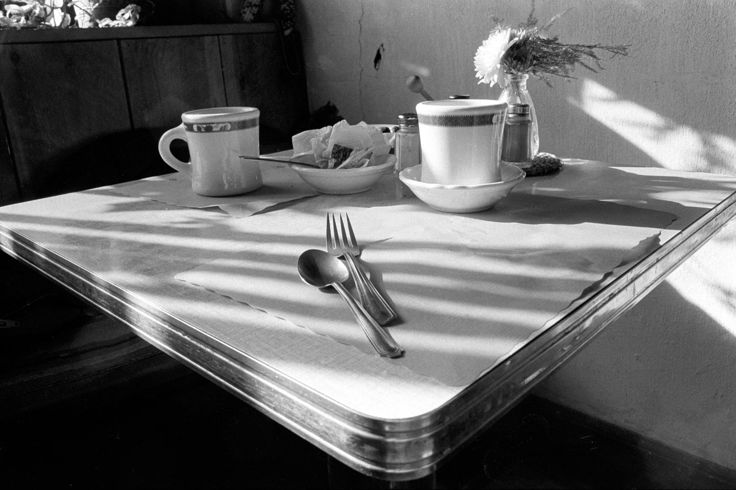 Place settings with silverware and white cups on a sunlit table.