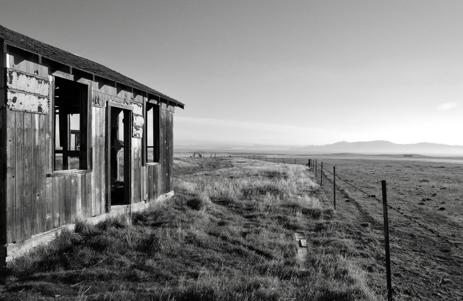 An old abandoned wooden shack sits isolated on a barren hillside against a cloudy sky.
