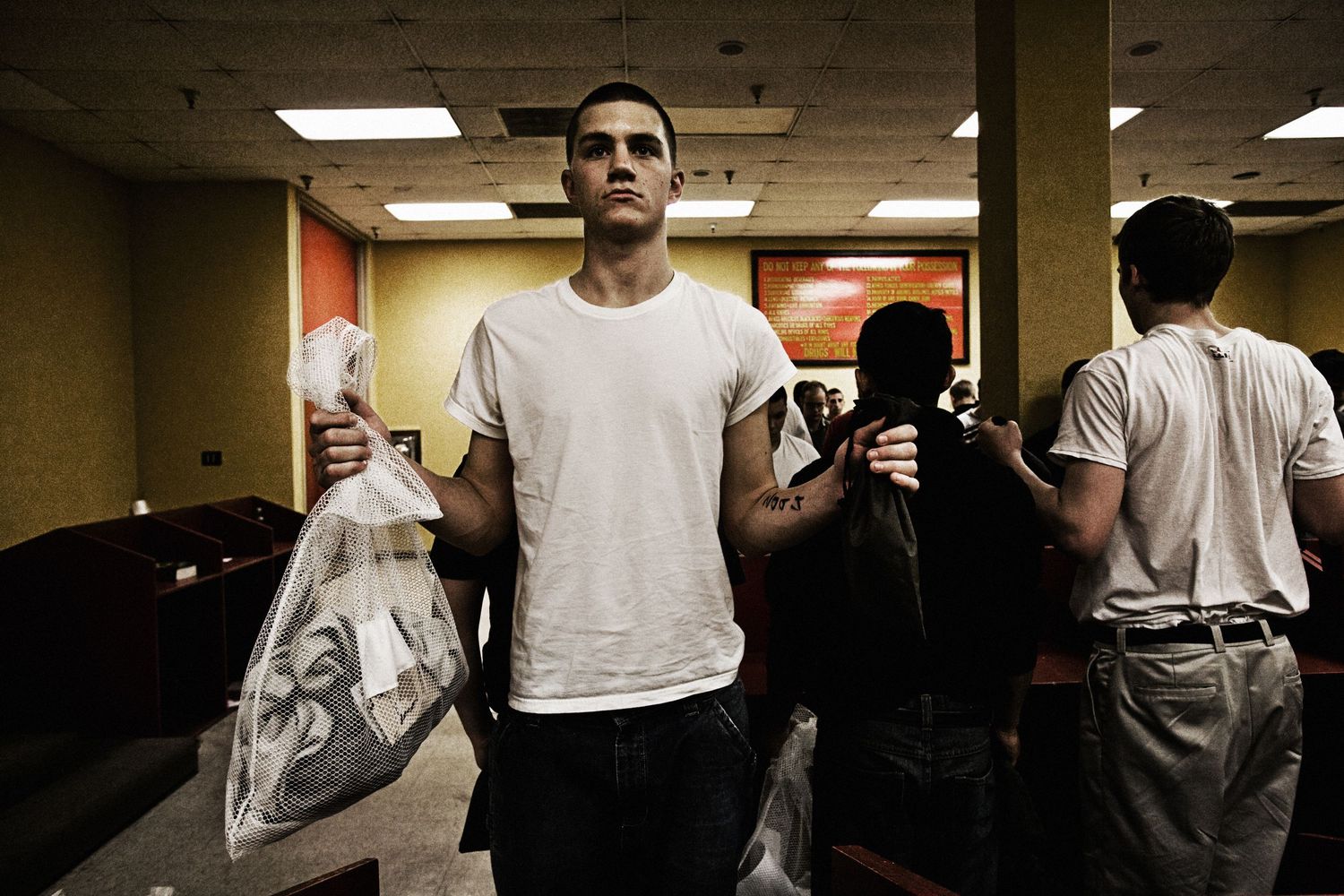 Military recruits standing in formation wearing white t-shirts in a dimly lit indoor space.