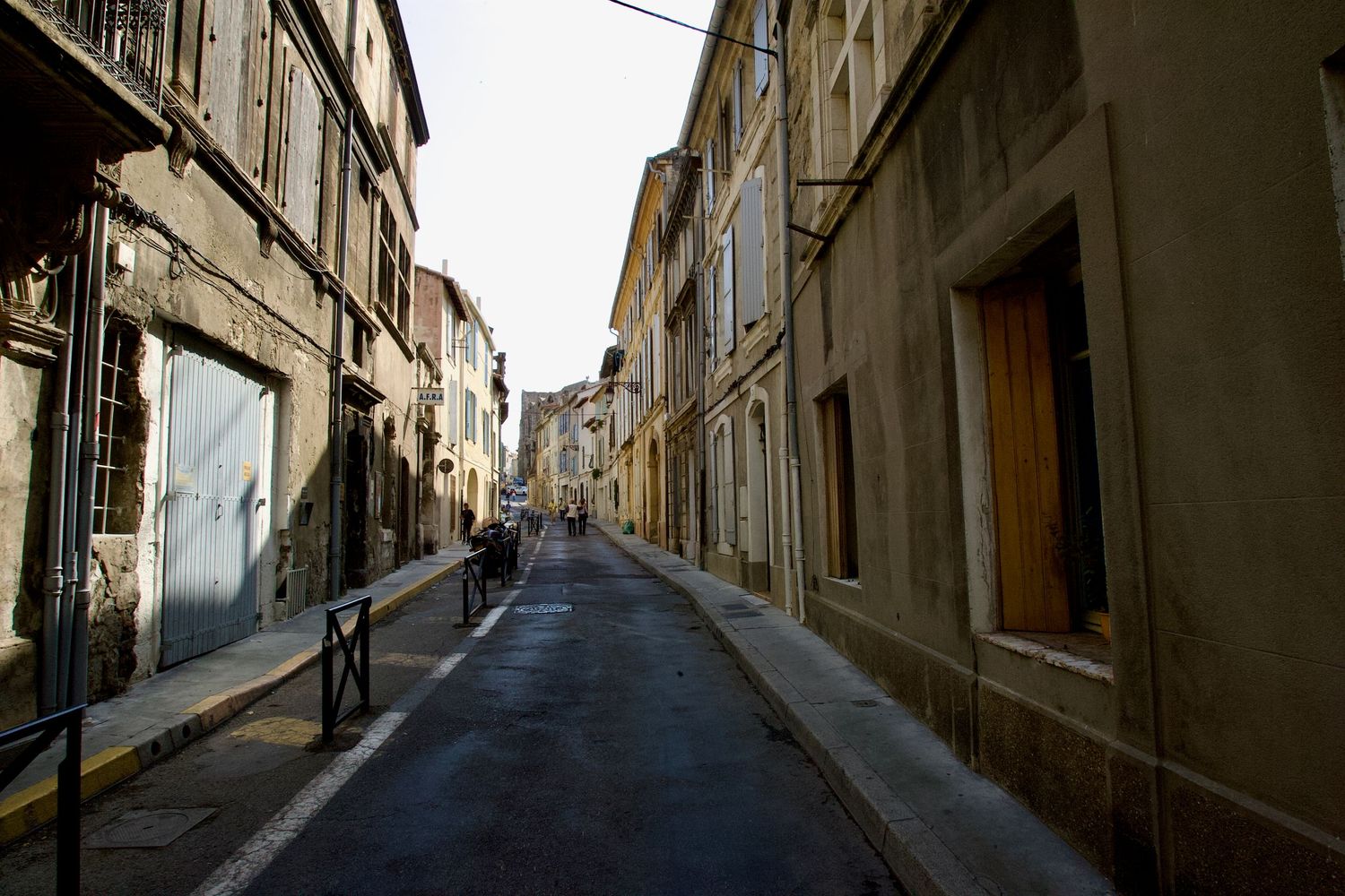 Historic stone buildings create shadowy corridors along narrow European street.