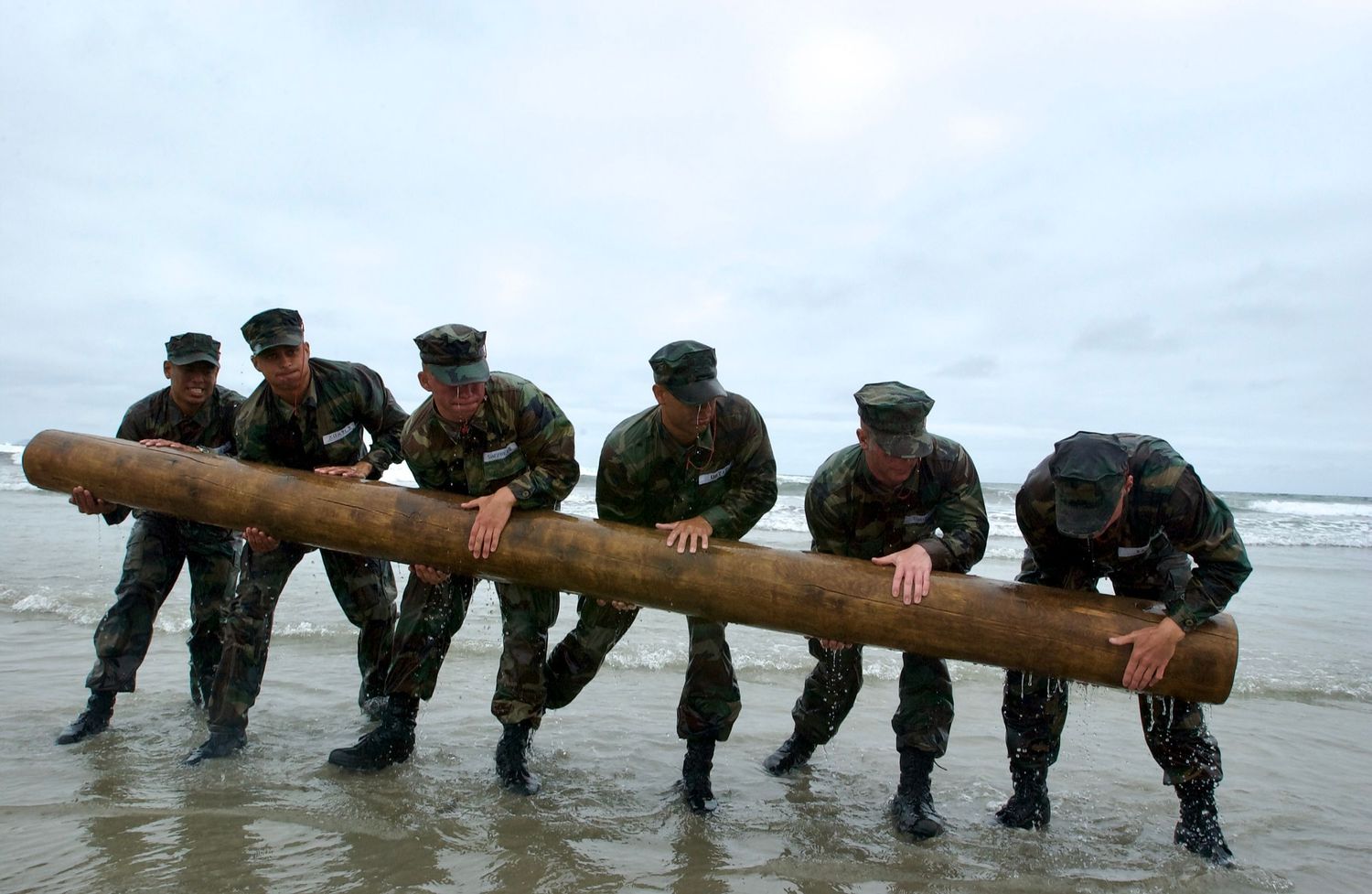 Military personnel work together to carry a large wooden log across a beach during training exercises.