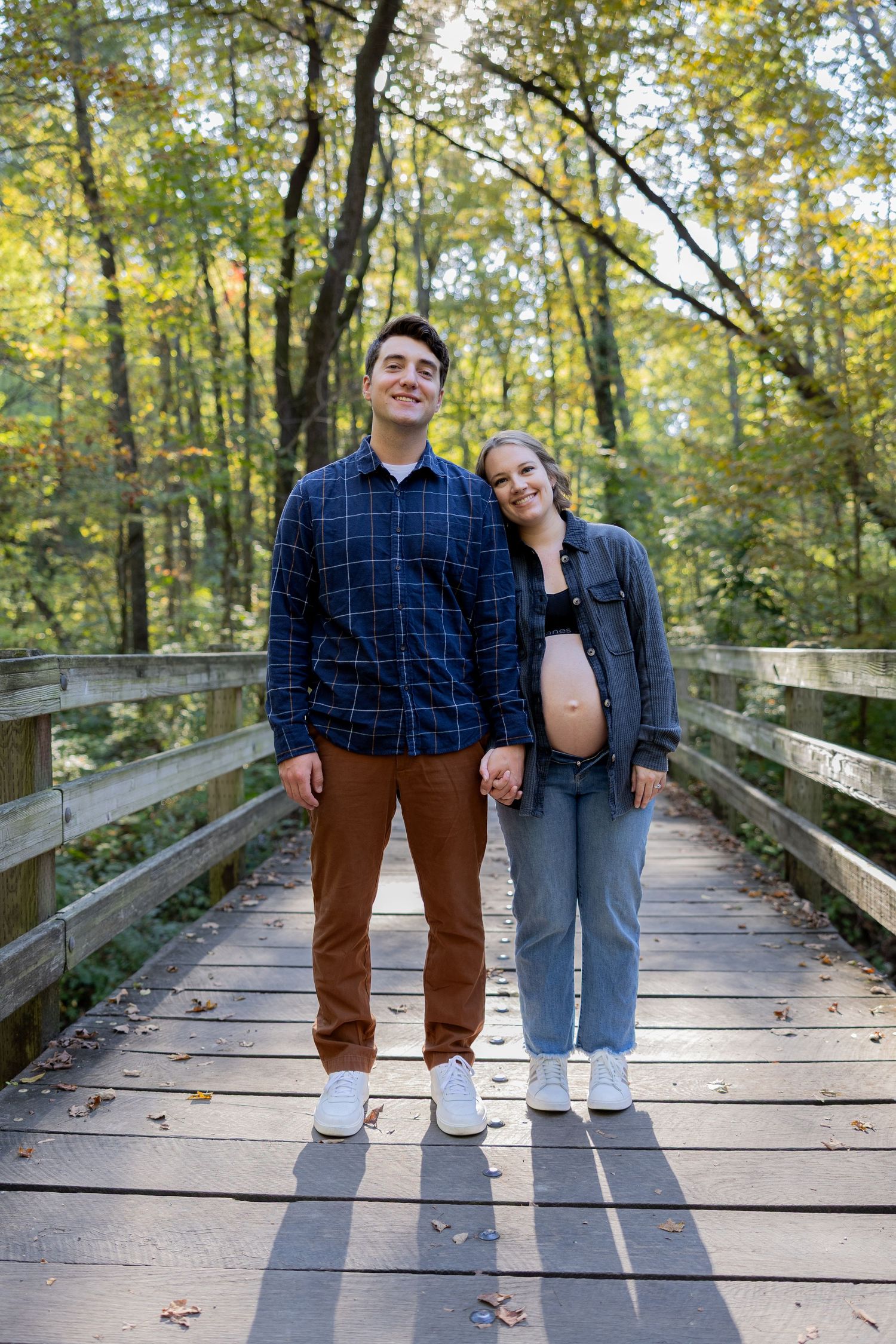 Couple stands together on wooden bridge in autumn forest setting during maternity photoshoot.