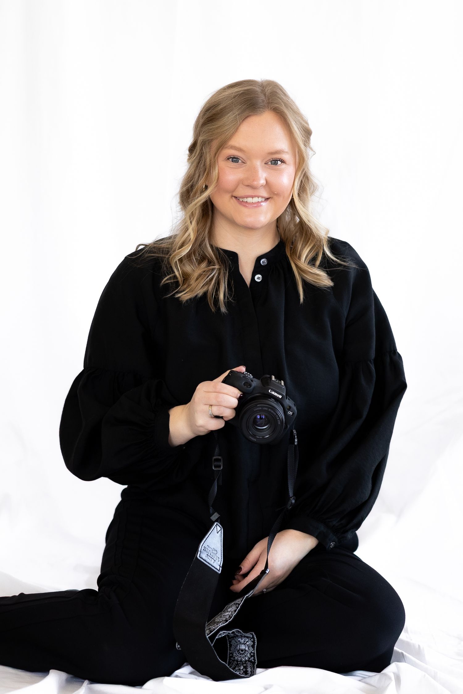 Professional photographer in all black clothing sits cross-legged while holding camera against white backdrop.