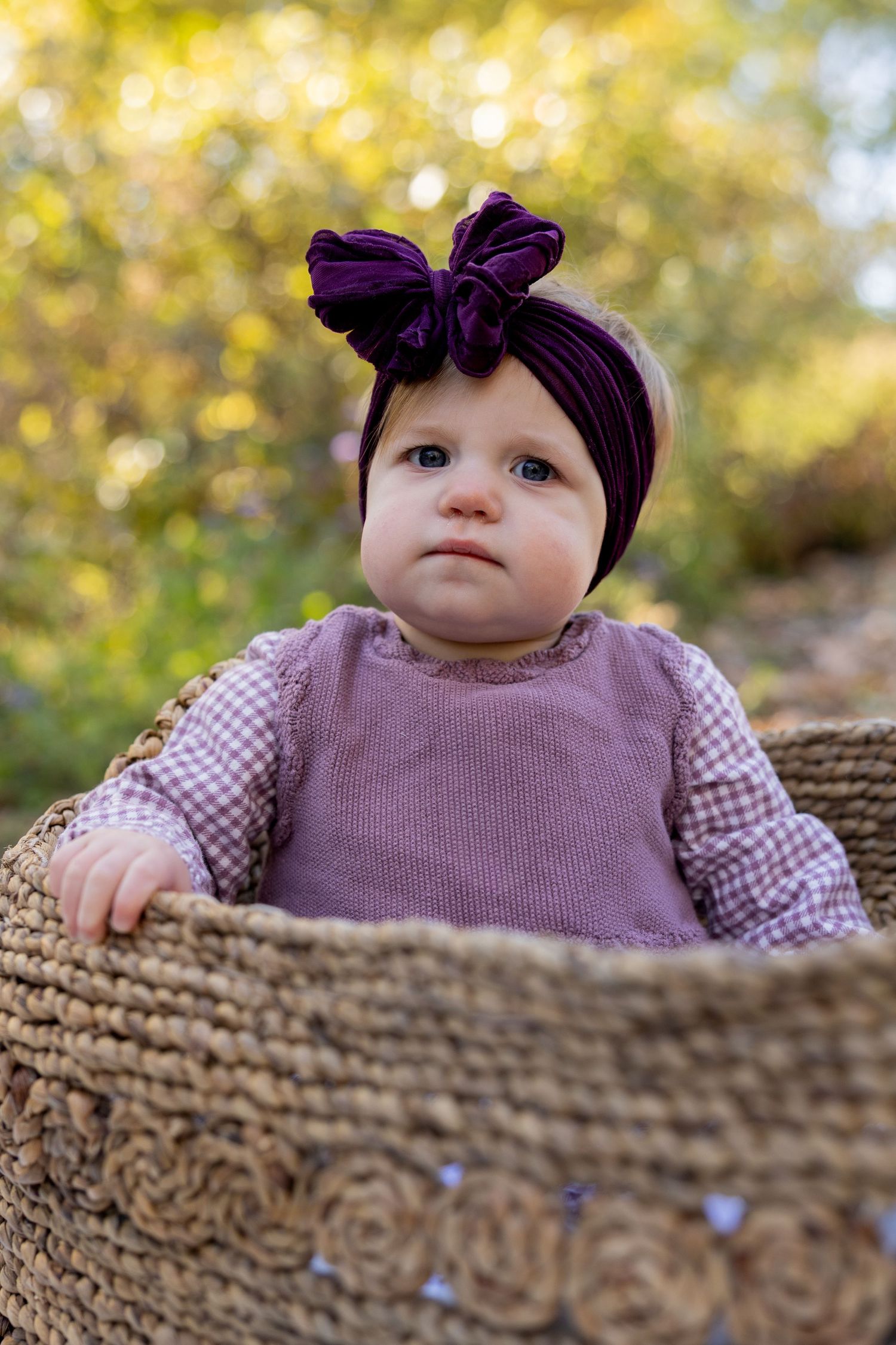 A baby wearing a purple dress and bow sits behind a textured brown wall in an outdoor autumn setting.