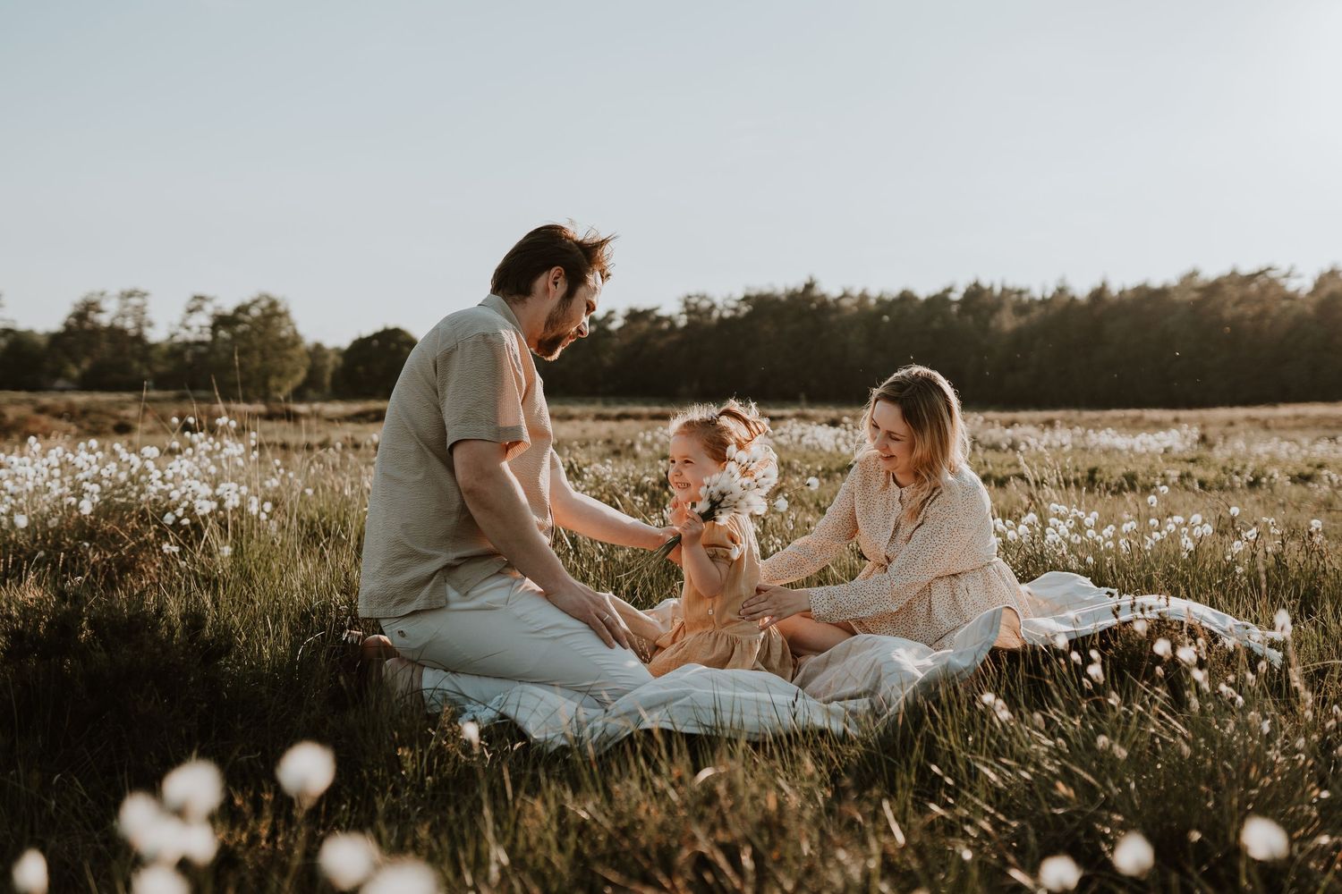 Family shares joyful moment on blanket in meadow filled with white flowers.