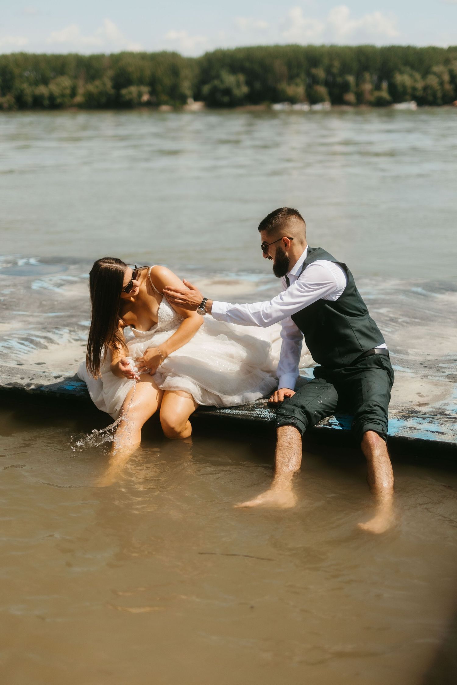 A couple in wedding attire playfully splash in shallow water at the edge of a lake on a sunny day.