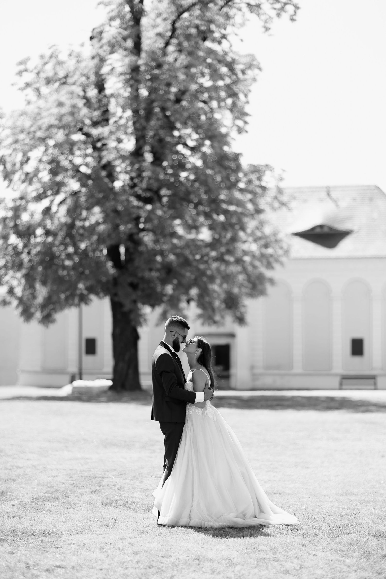 A black and white wedding photo shows a couple embracing on a lawn with a grand building and tree in the background.
