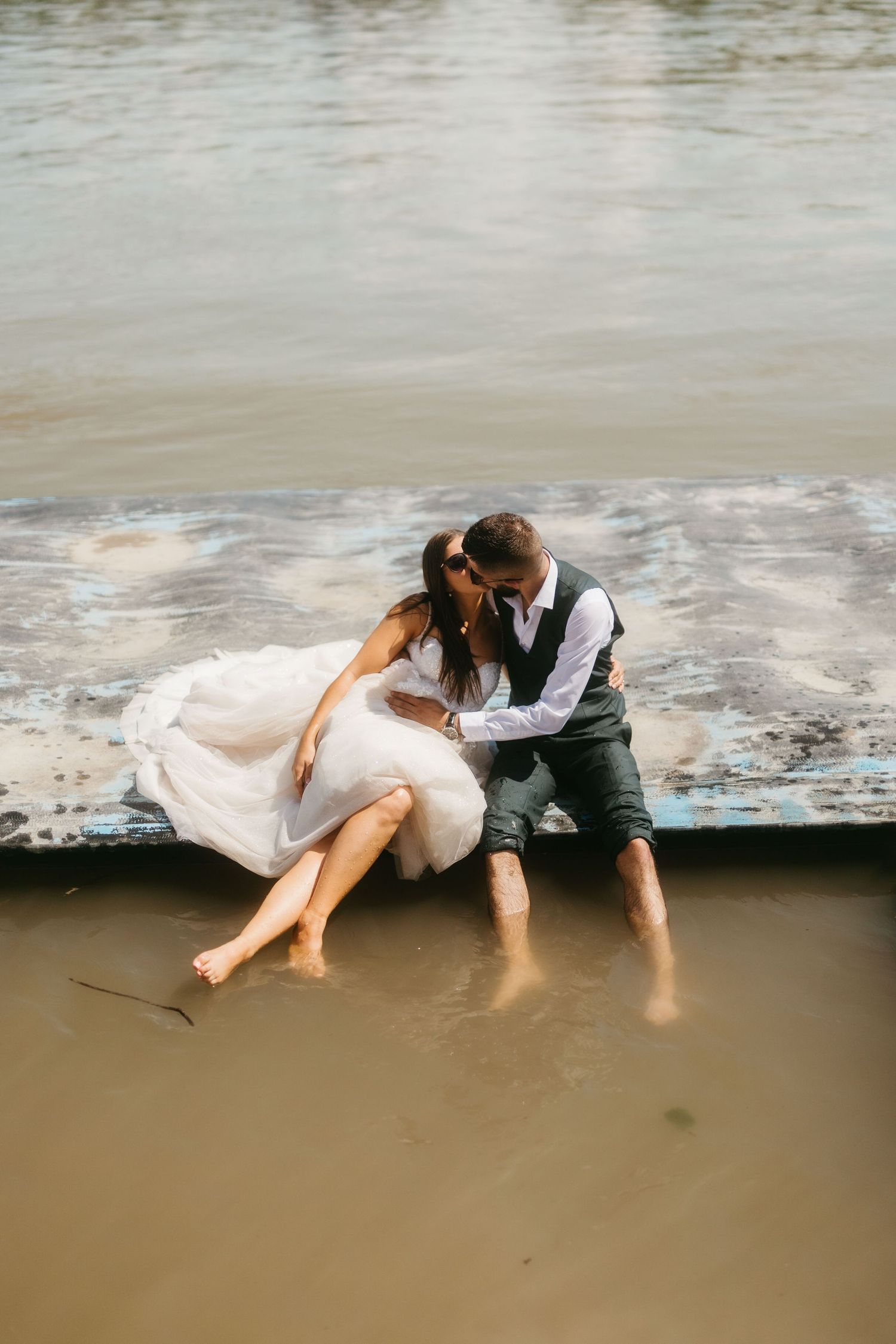 Two people in wedding attire sit together with feet in shallow water during a romantic photoshoot by the shore.