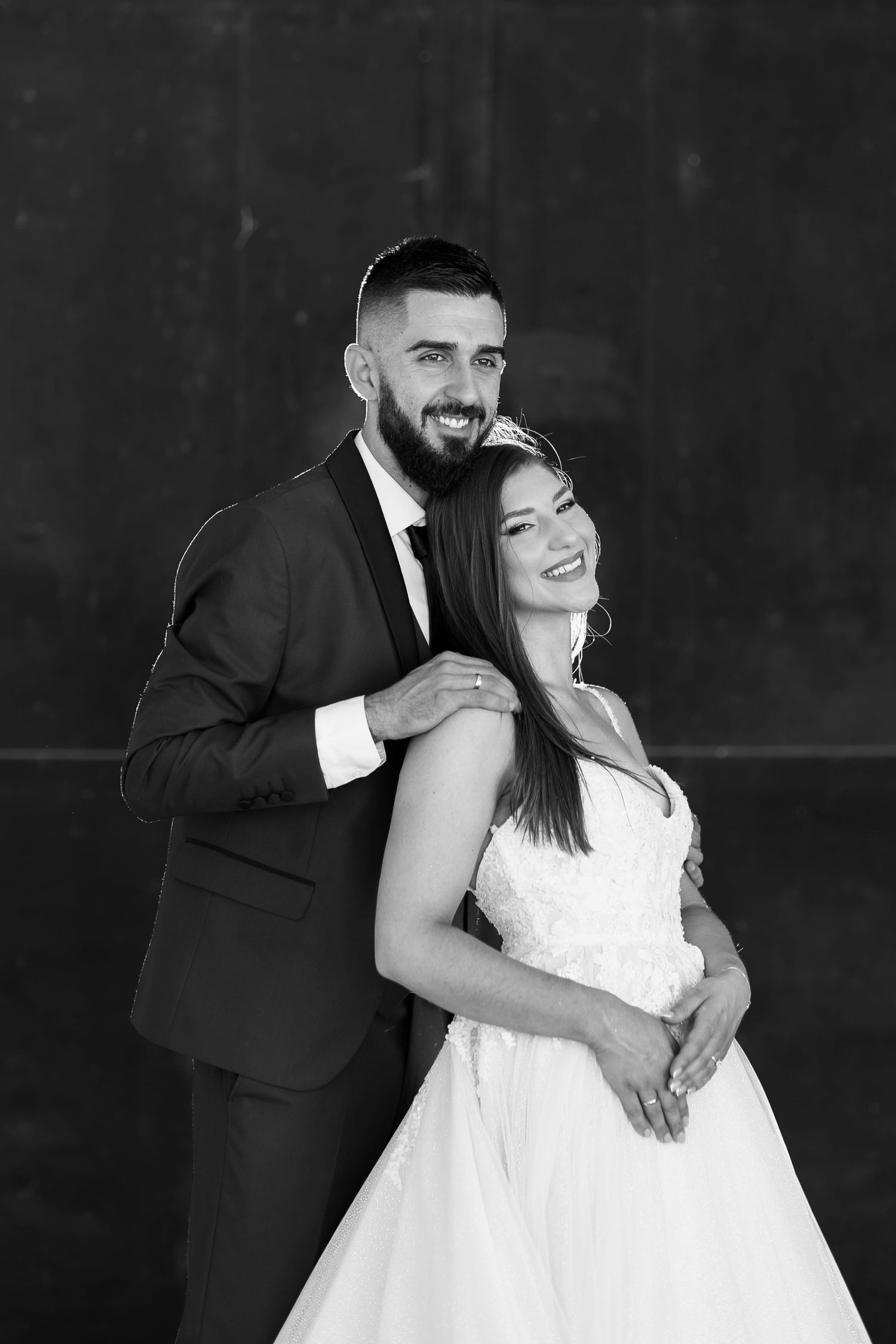 Black and white portrait of a couple in formal wedding attire embracing against a dark background.