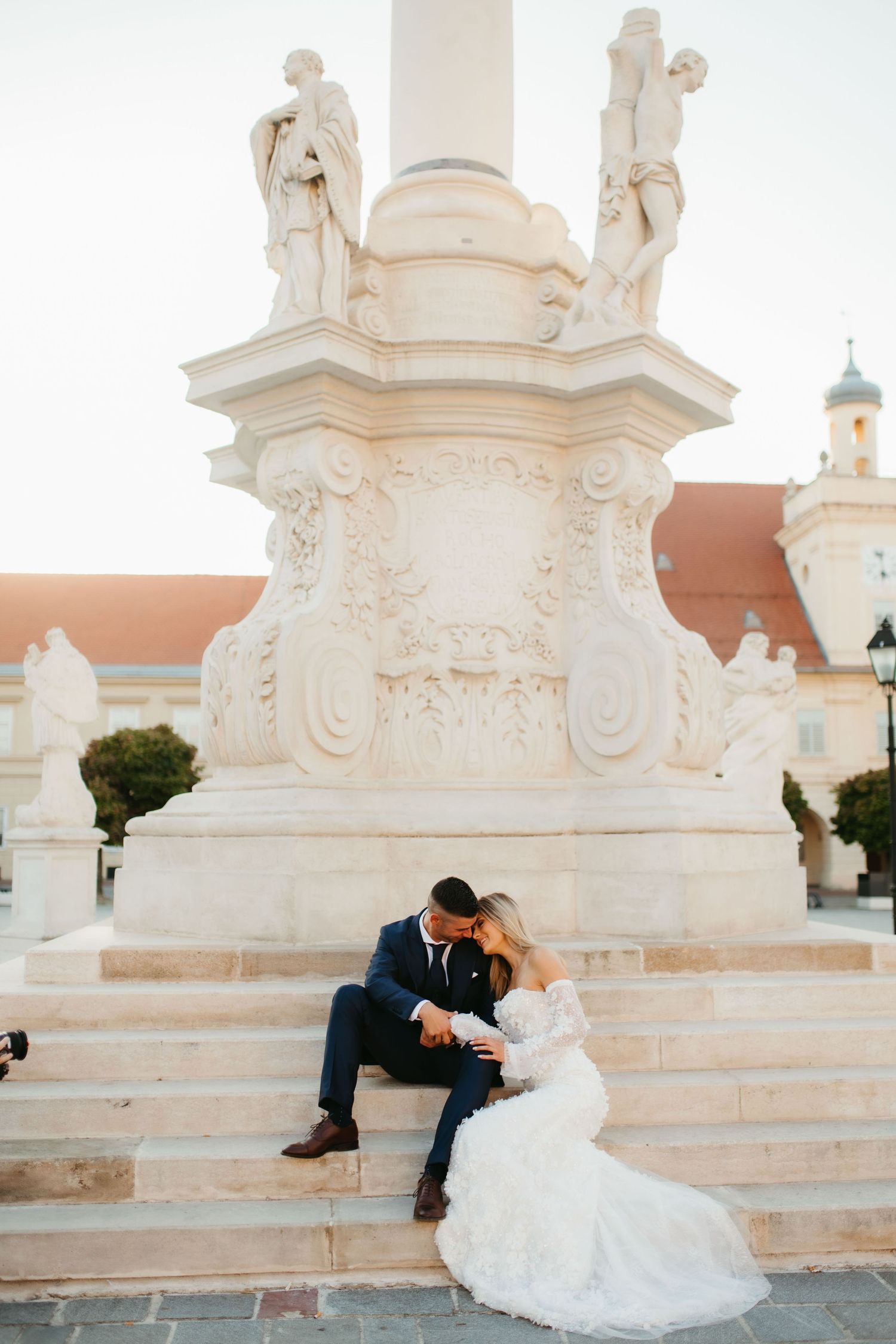A wedding couple sits intimately on marble steps near a decorative stone monument.