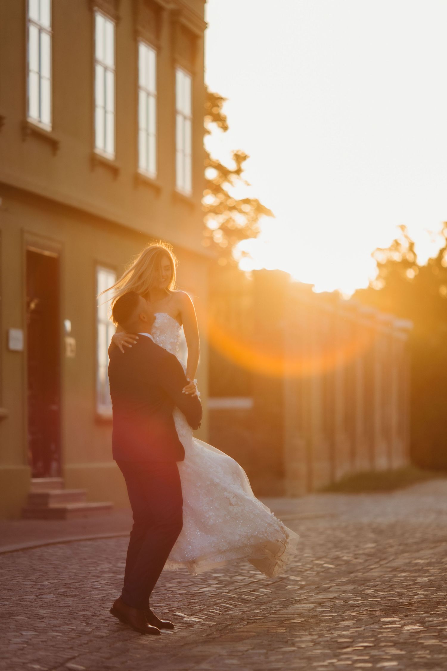 A couple in wedding attire dance on cobblestones during a warm sunset.