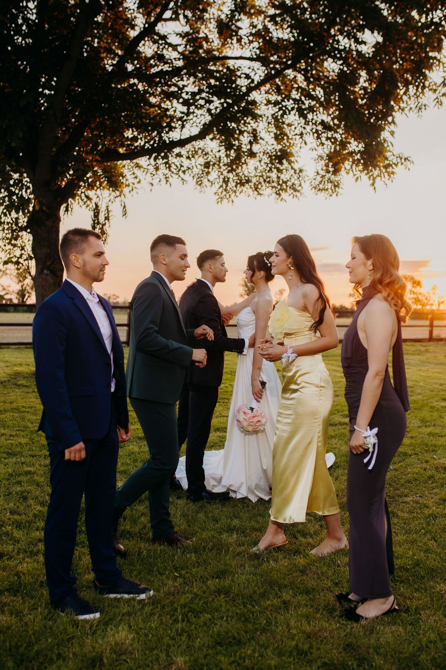 Wedding guests gather on a lawn at sunset under a large tree during an elegant outdoor celebration.