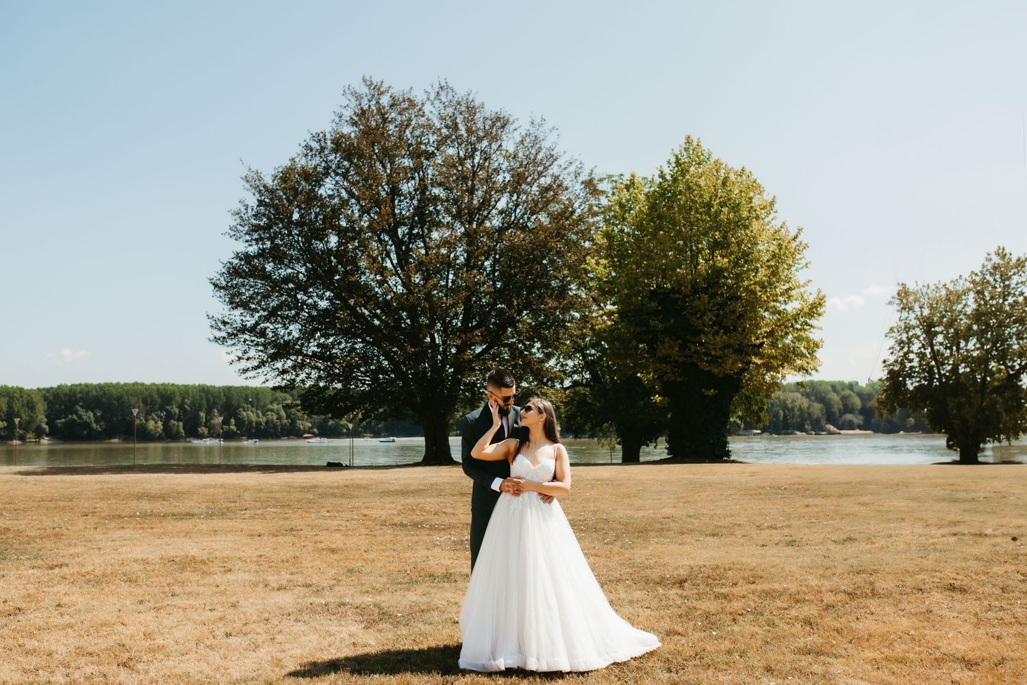 A newlywed couple embraces in a sun-drenched field with large trees and a scenic landscape in the background.