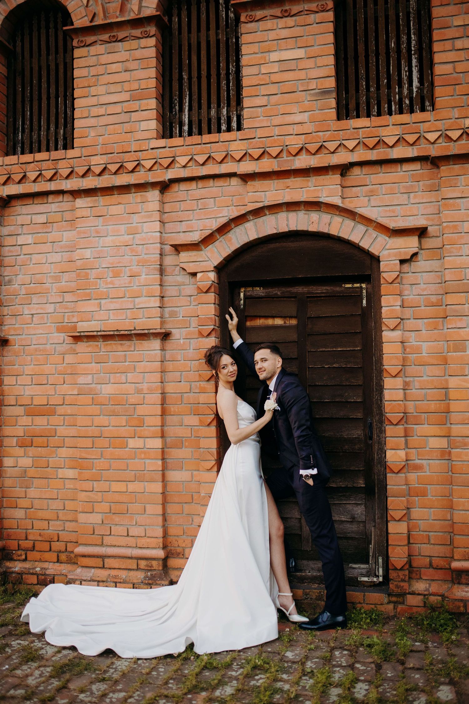 A wedding couple poses against a rustic brick wall with arched doorway and flowing white dress in elegant outdoor setting.