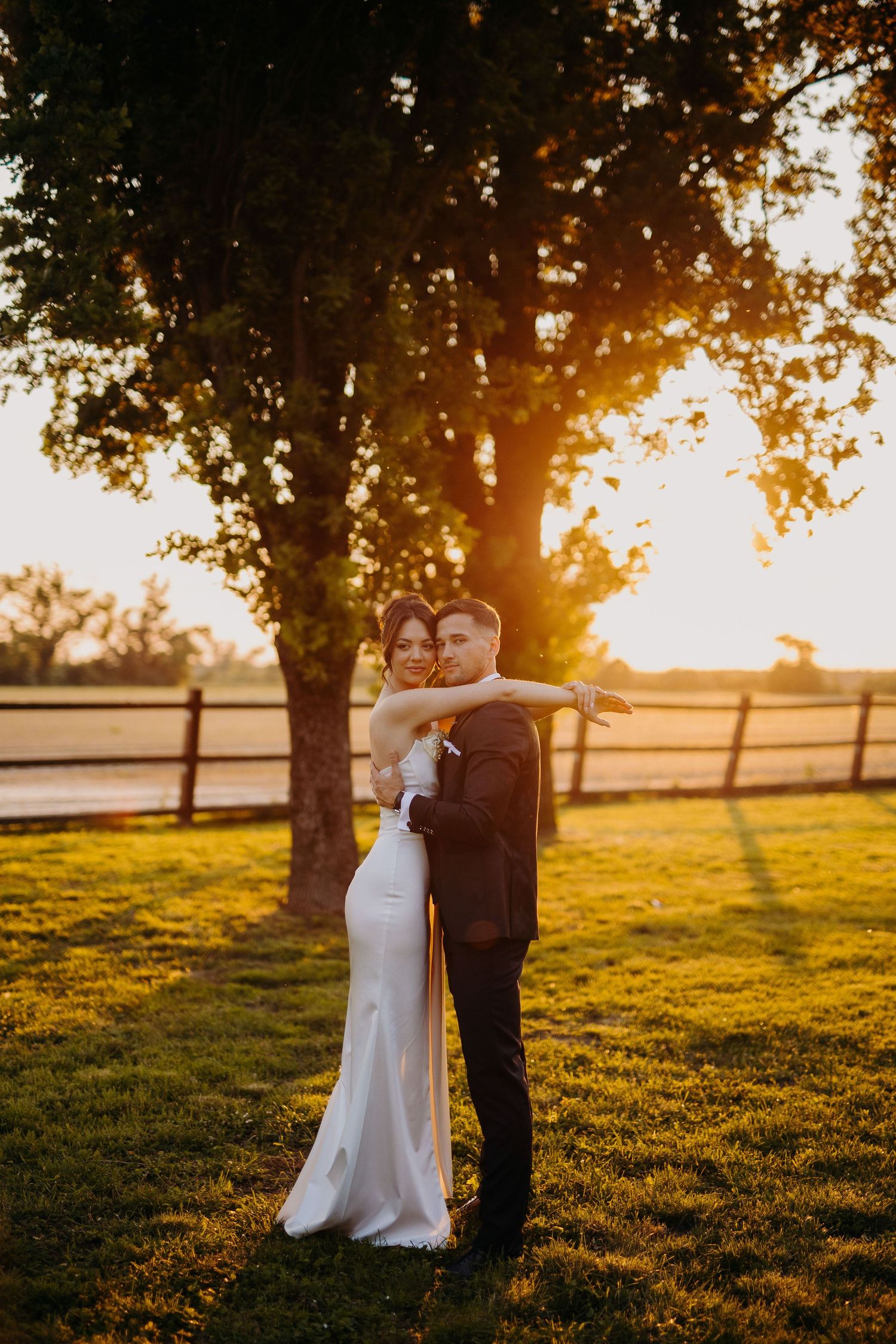 A romantic golden hour wedding portrait with newlyweds embracing in a sunlit field with wooden fences and trees.