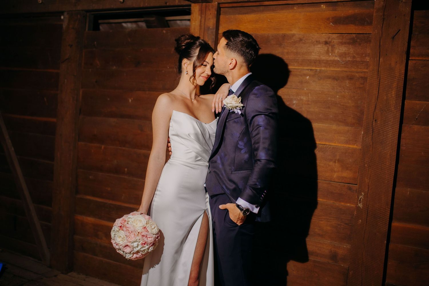 An intimate moment between newlyweds sharing a romantic embrace in a rustic wooden barn-style venue.