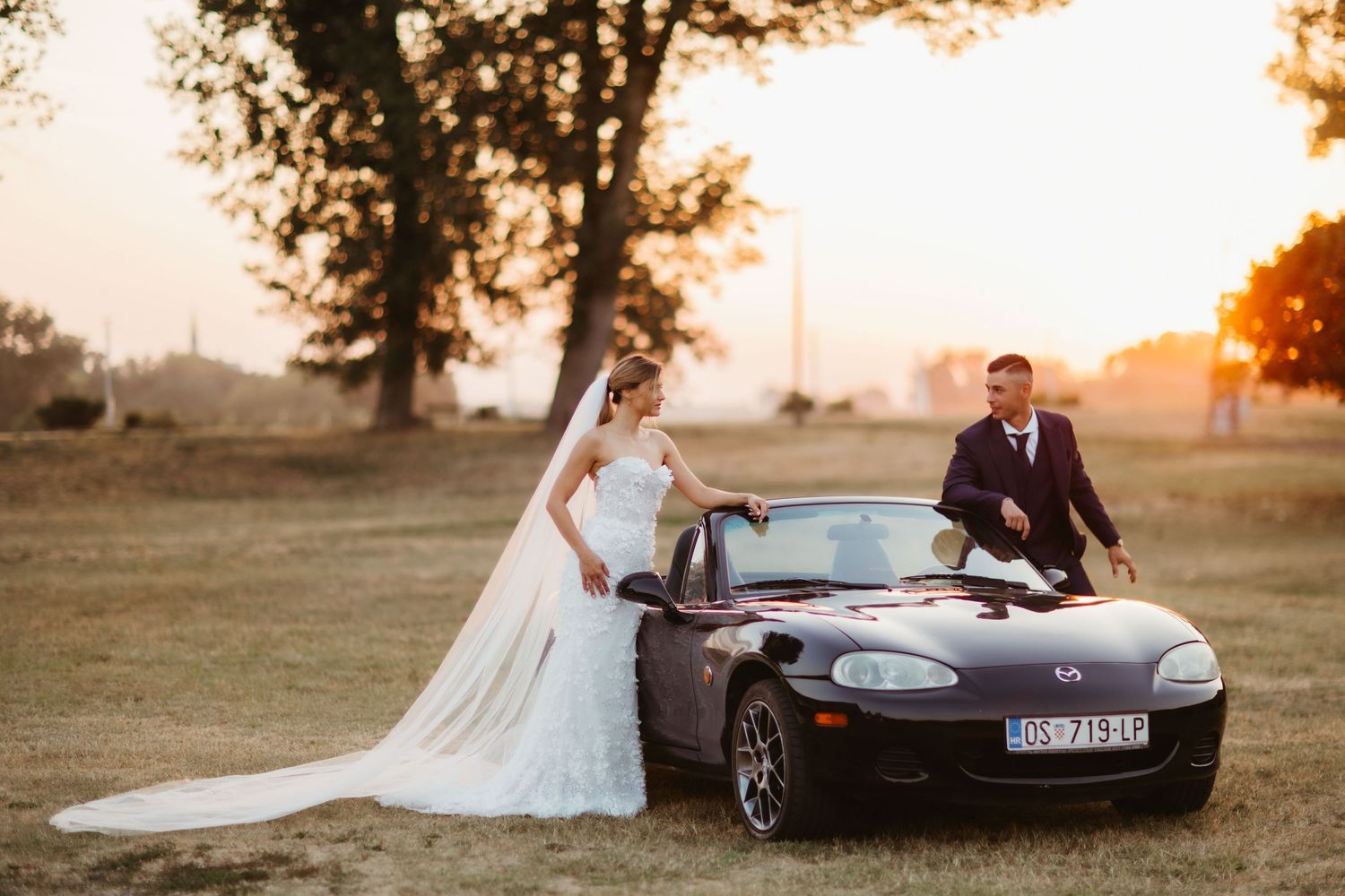 Newlyweds pose with a black sports car in a field during golden hour.