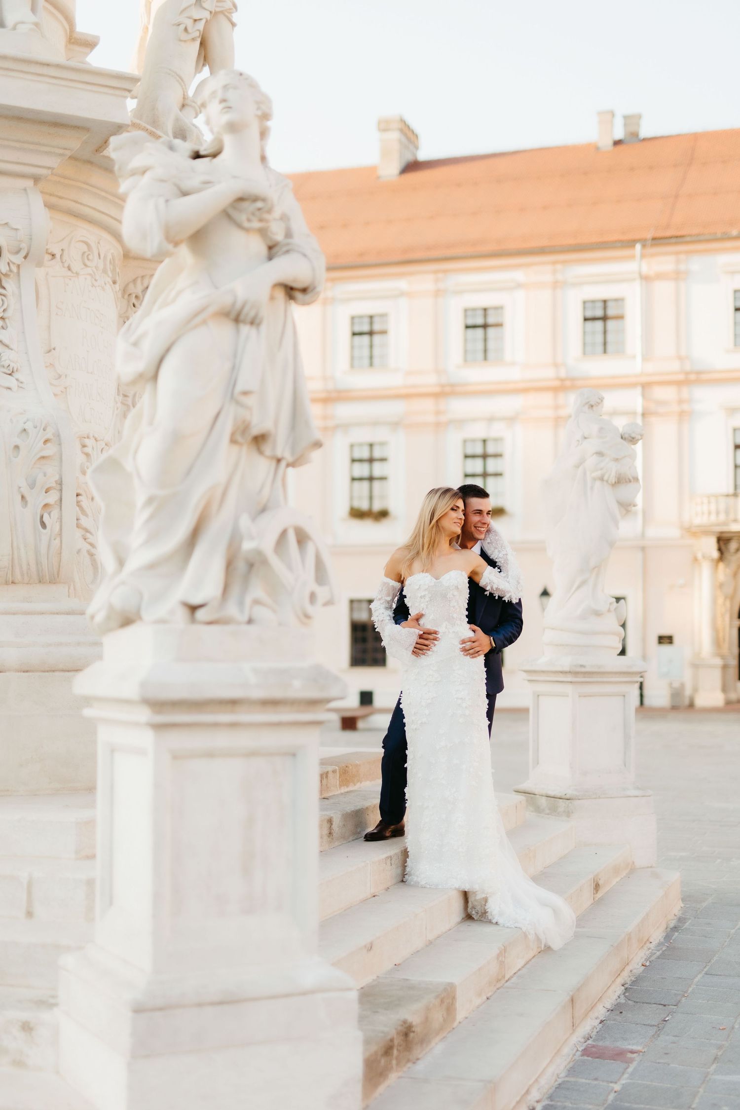 Wedding couple poses on stone steps with classical statues and architecture.