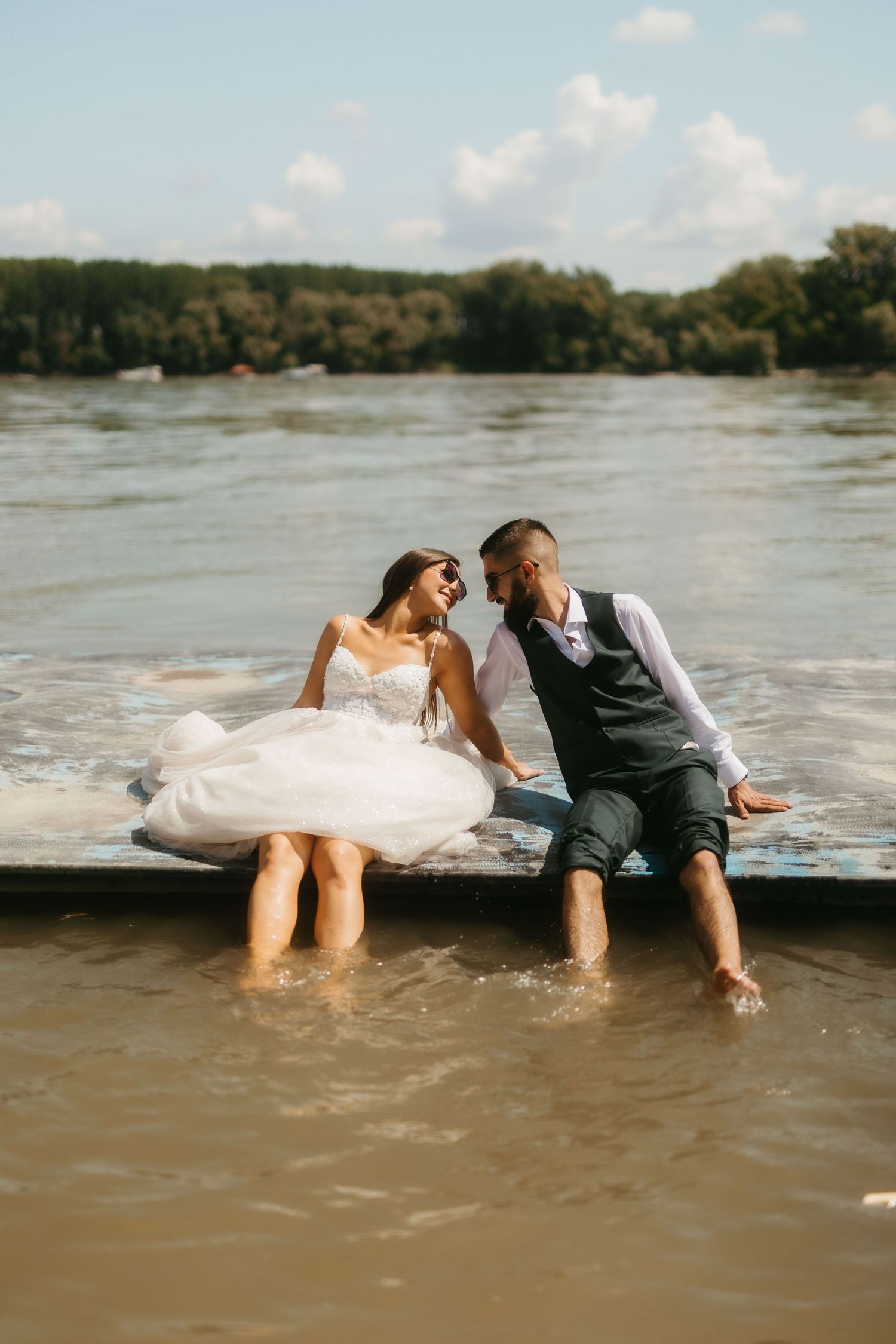 A couple sits and splashes their feet in a river from a dock, wearing formal wedding attire on a sunny day.