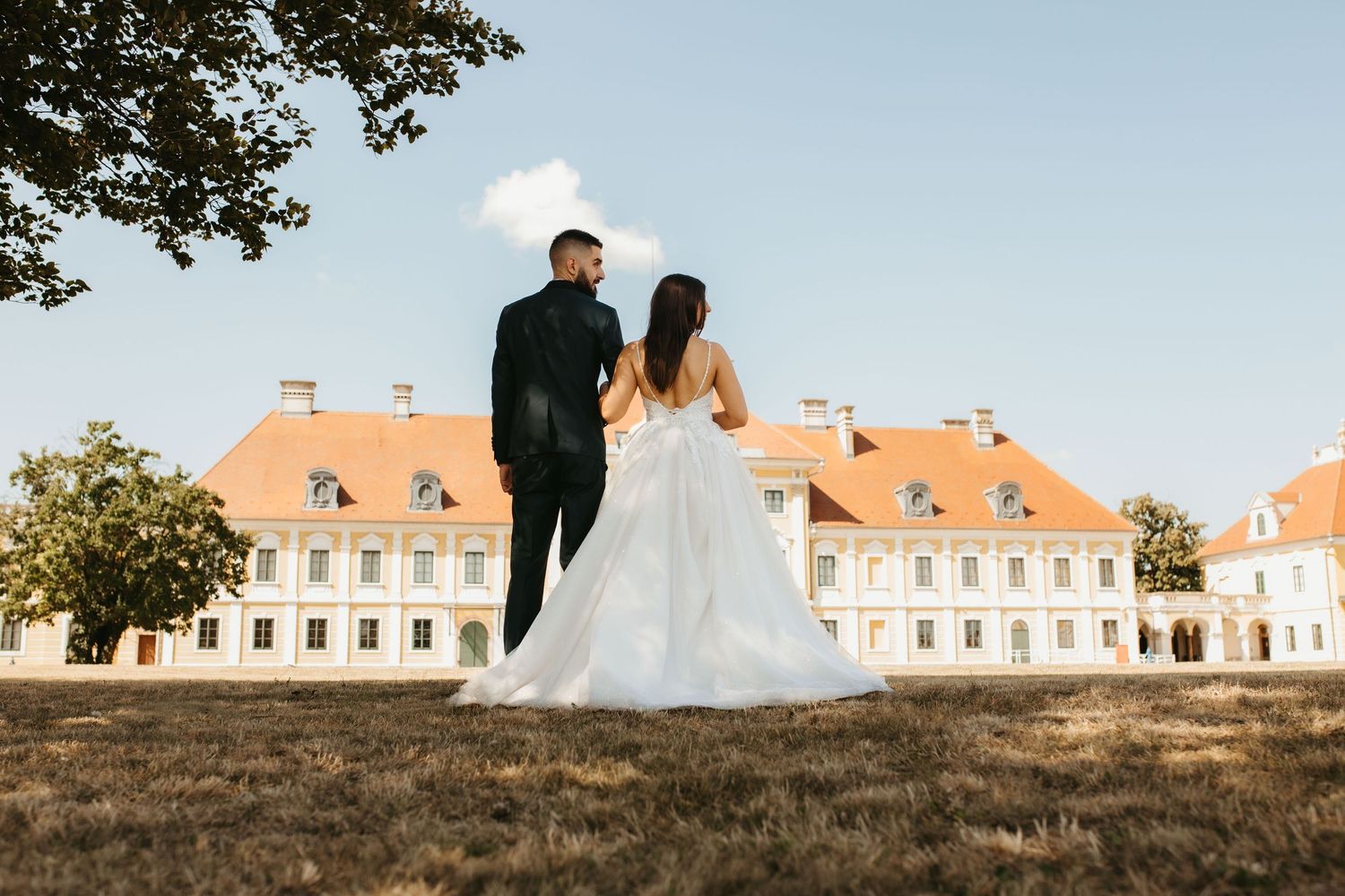 Wedding couple poses on a grassy hill in front of a grand white baroque mansion on a sunny afternoon.