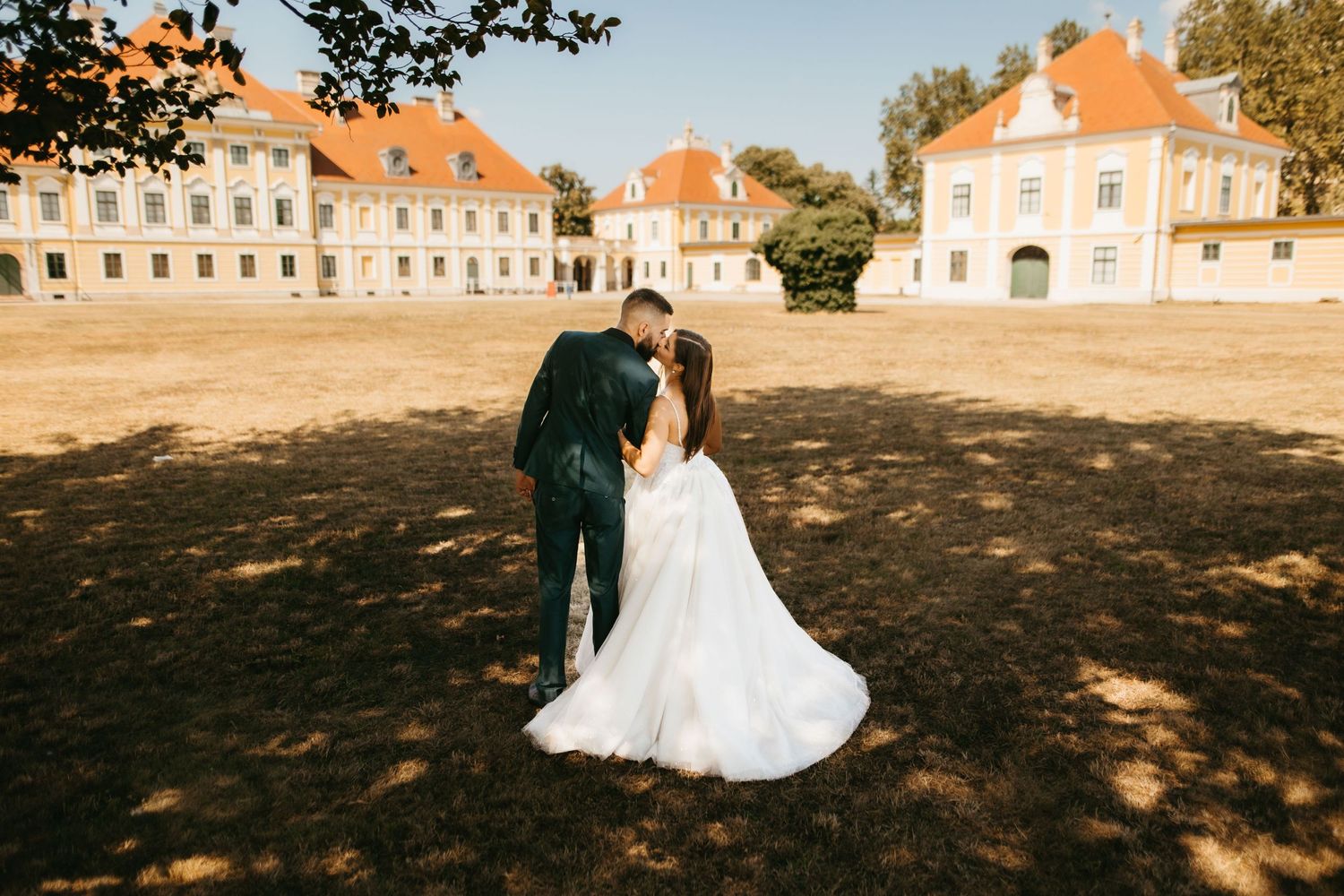 A romantic couple in wedding attire stands before historic orange-roofed buildings on a sun-dappled courtyard.