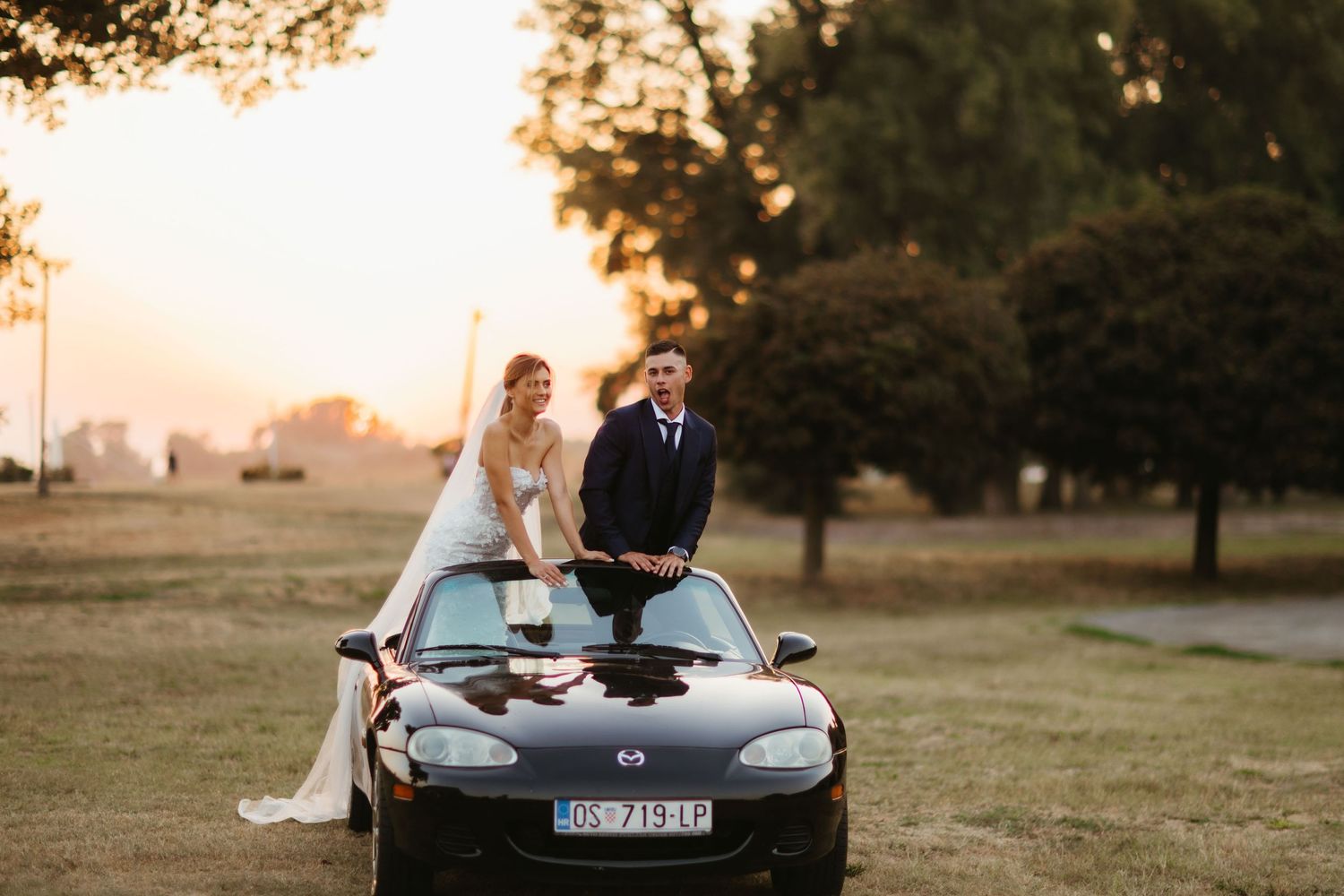 Couple celebrating their wedding day standing through sunroof of black Mazda Miata at sunset.