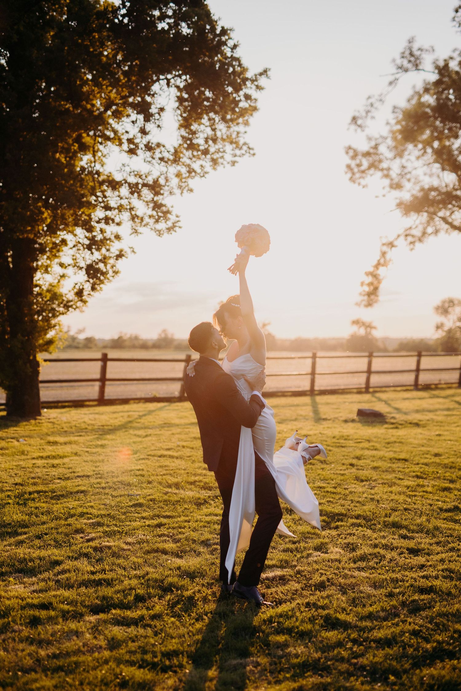 Silhouetted couple embraces in a golden sunset field with wooden fence and trees casting long shadows.