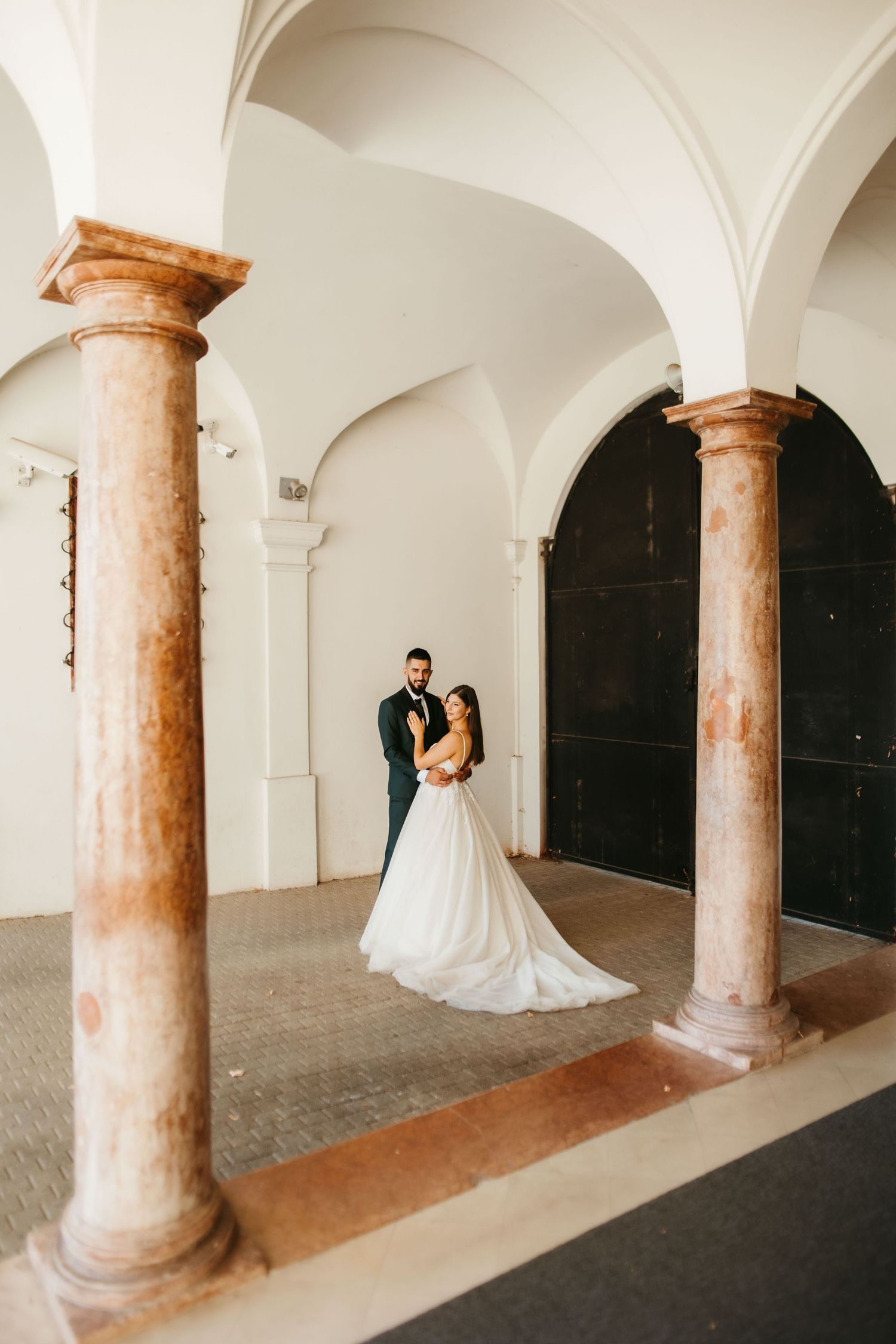 A romantic wedding photo under ornate marble columns and arches with elegant architectural details.
