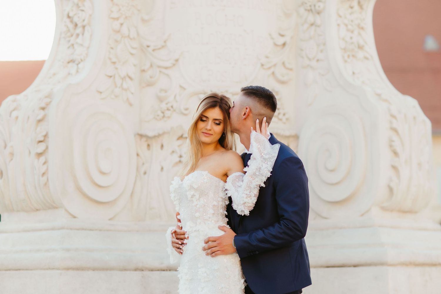 Newlyweds share an intimate moment against an elegant baroque architectural backdrop.