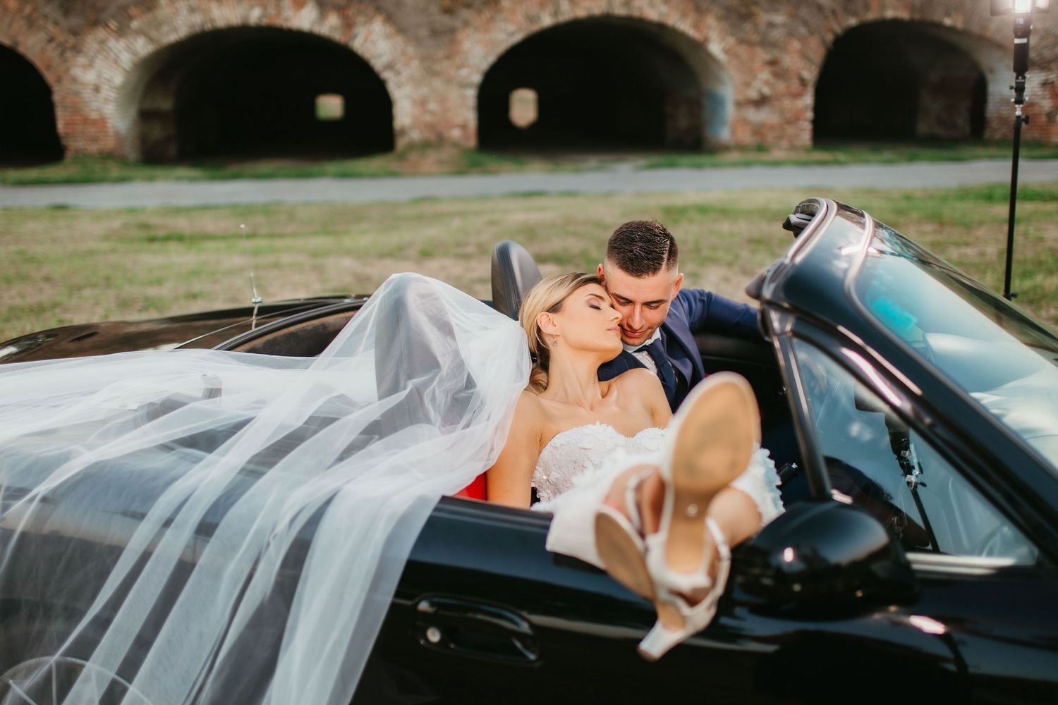 Newlyweds share an intimate moment in a convertible against a brick archway backdrop.