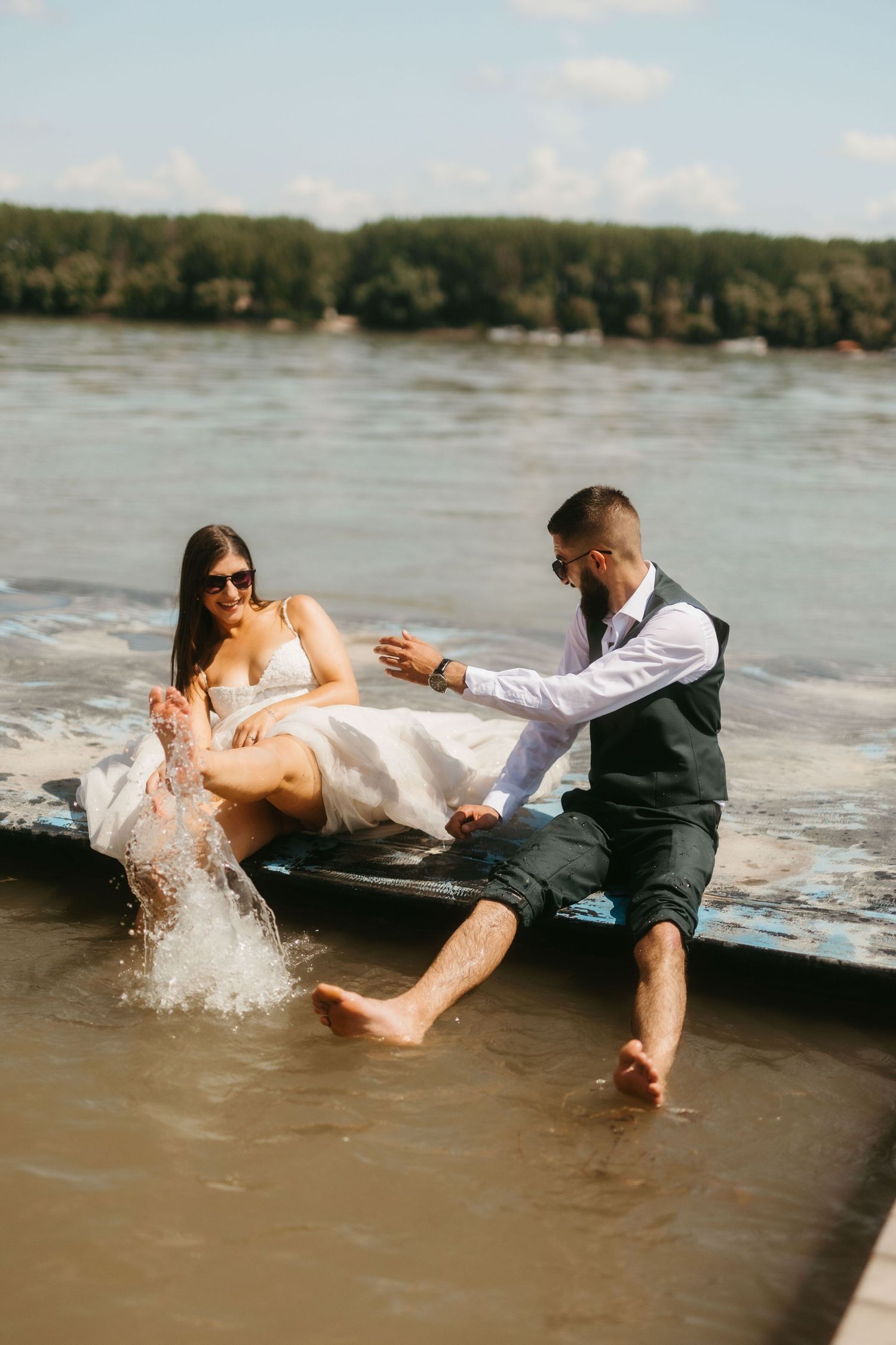A couple in wedding attire playfully splashing in the water during a lakeside photo session.