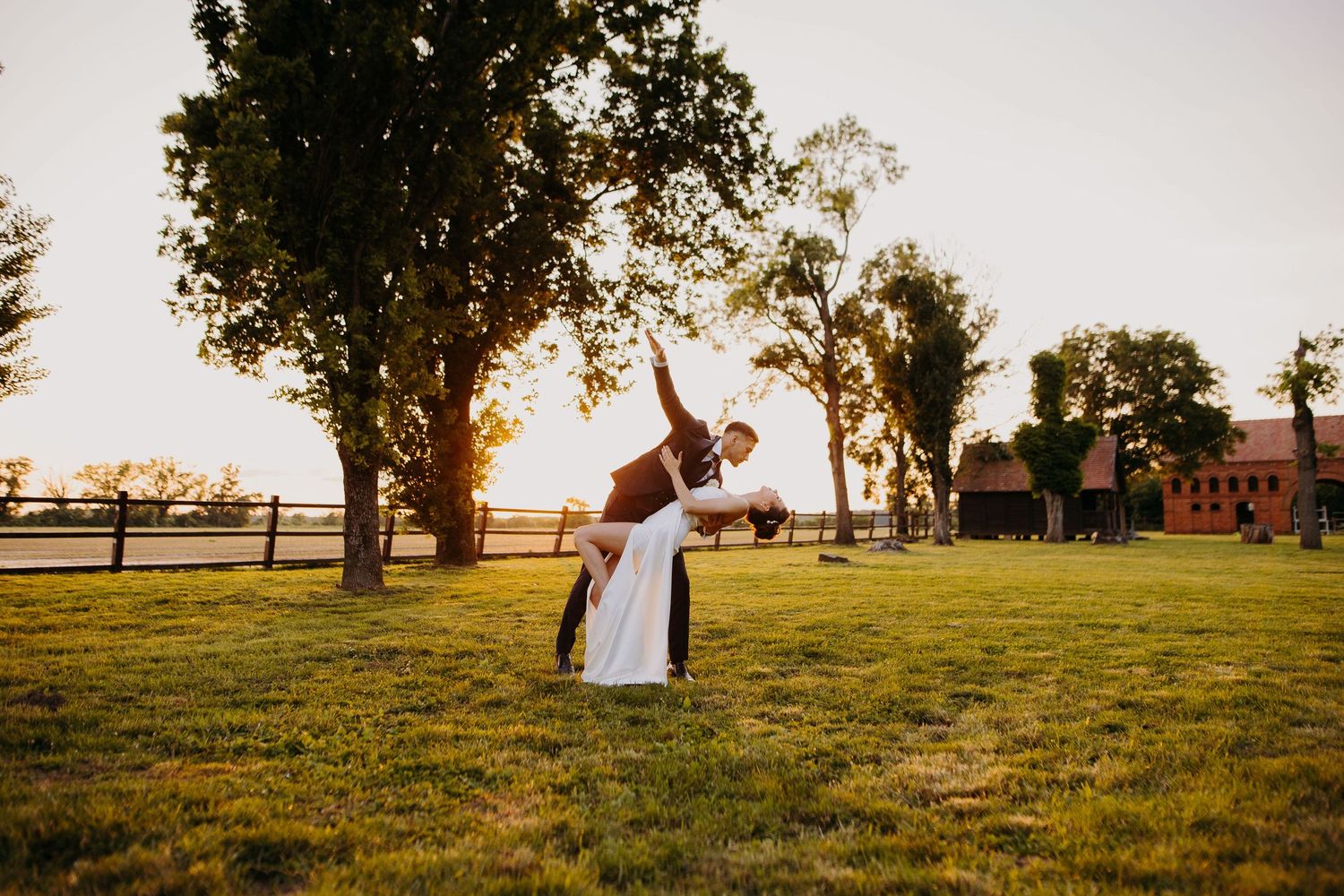 A couple shares a romantic dip during sunset in a rustic farm field with trees and fencing in the background.