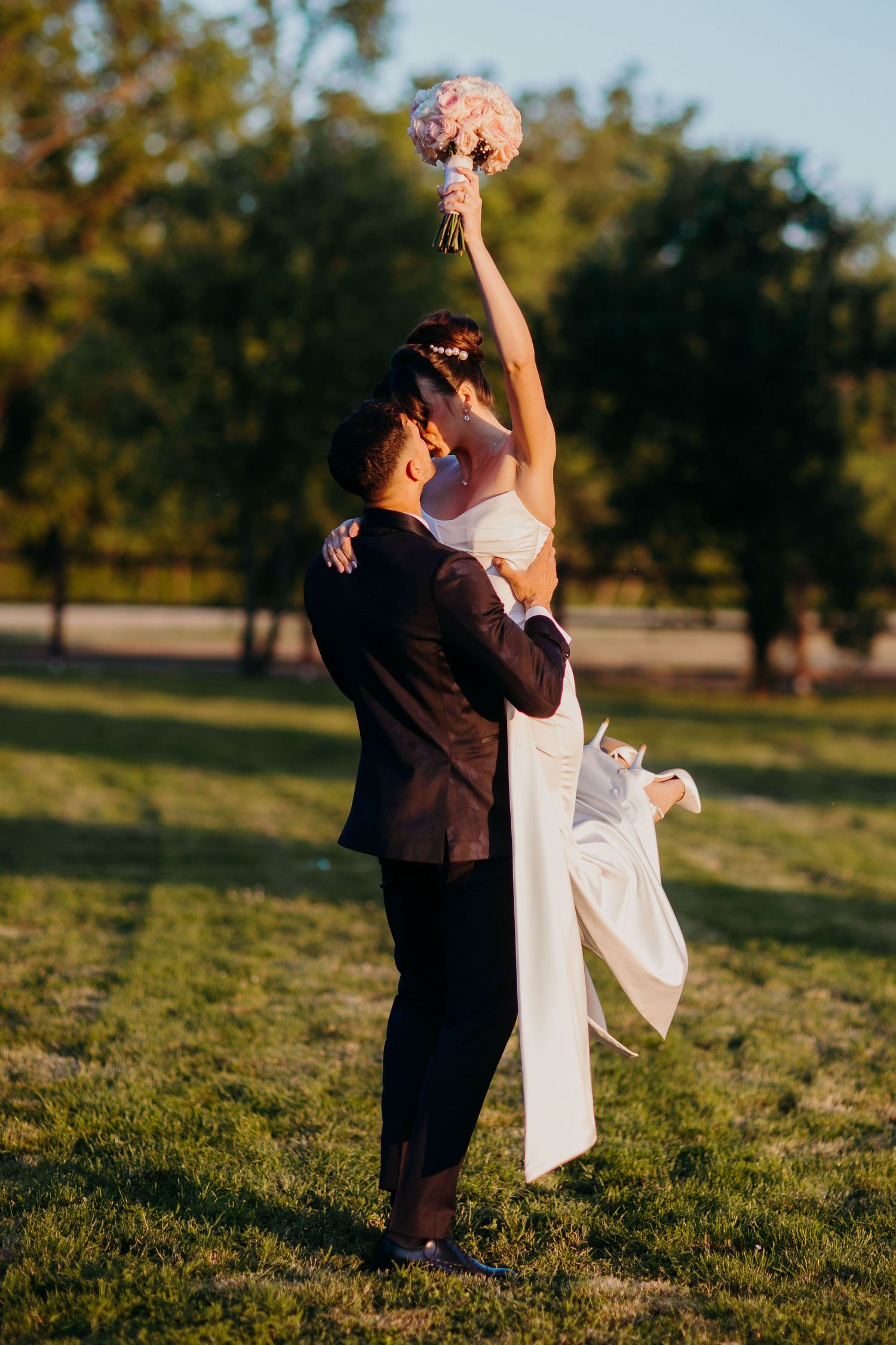 A wedding couple strikes a dramatic dance pose outdoors with a pink bouquet held high against a natural backdrop.