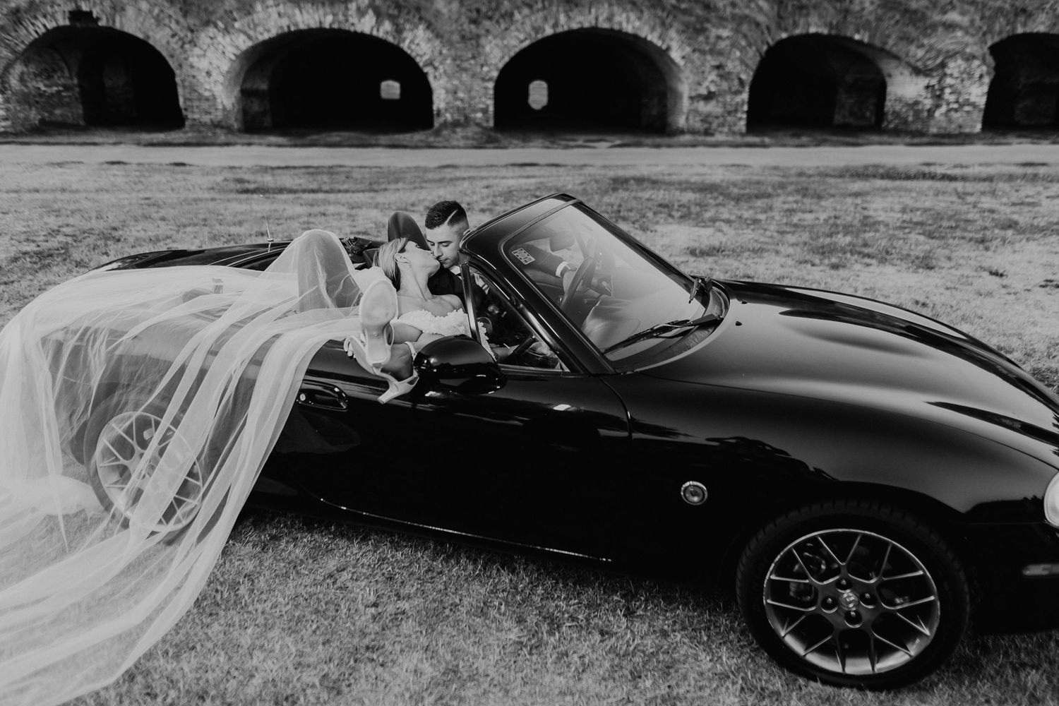 Black and white photo of a wedding couple in a classic sports car by stone arches.
