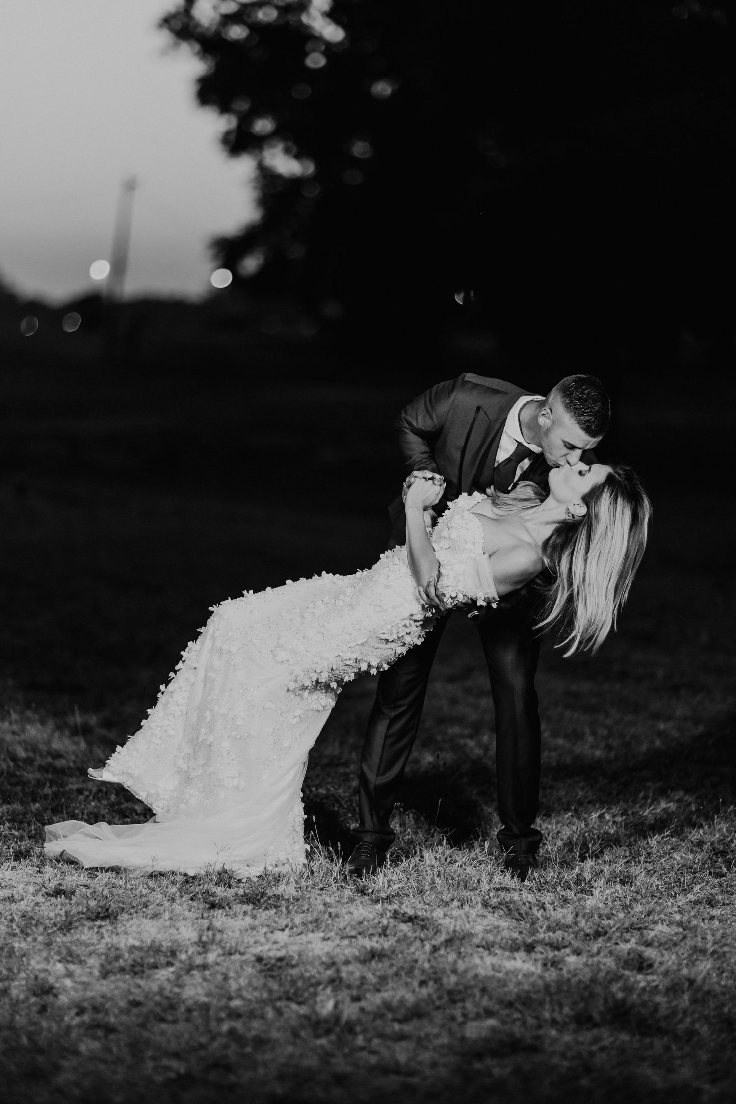 A black and white wedding photo captures a romantic dip pose against a dusky evening sky.