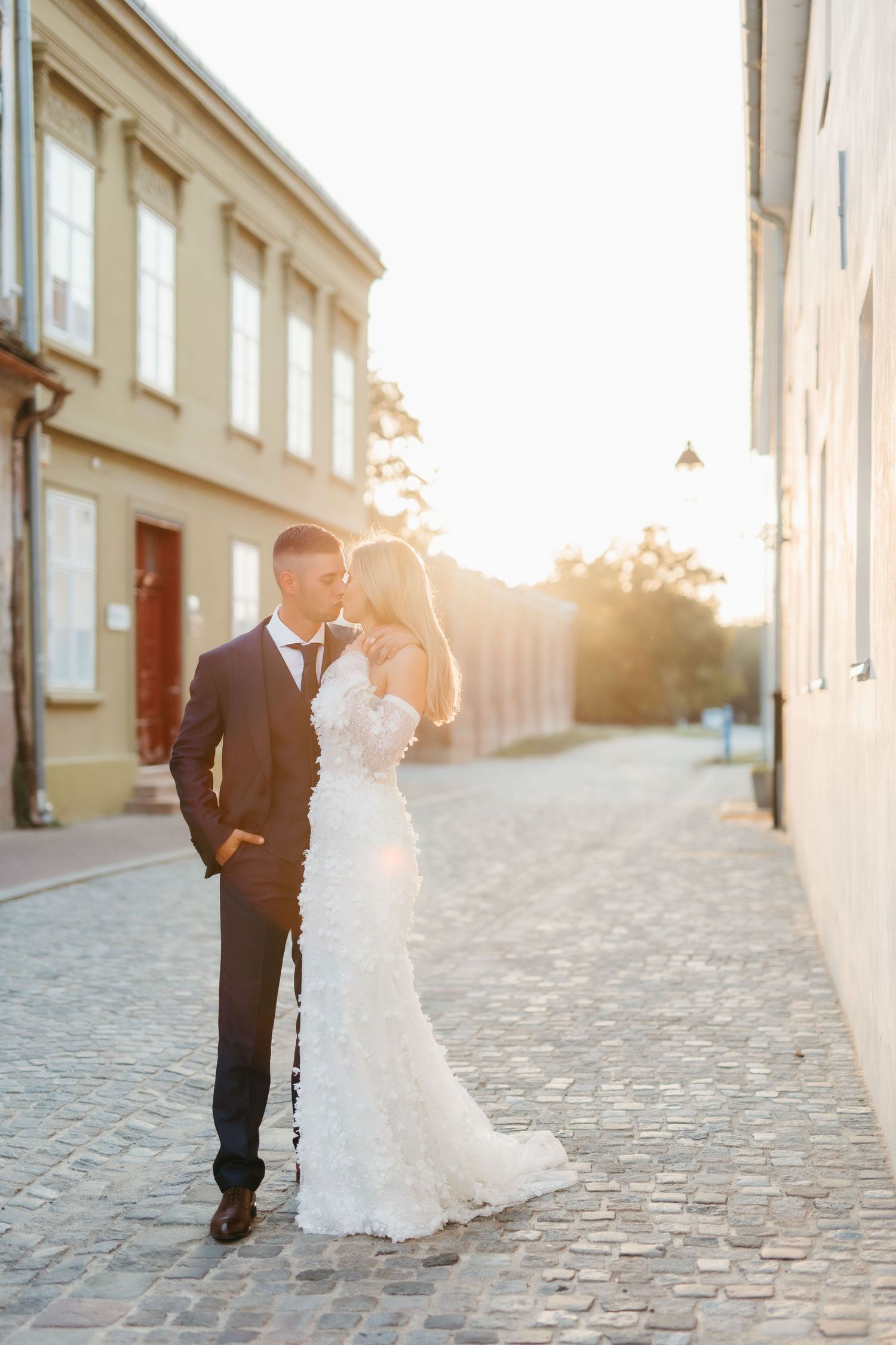 Wedding couple poses on cobblestone street with sunlit buildings in background.