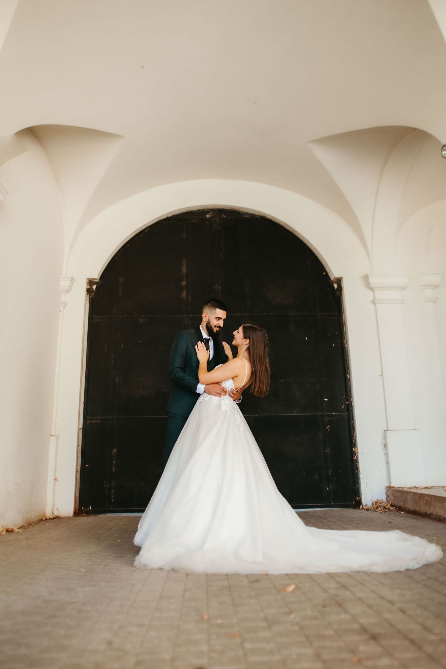 A romantic wedding couple embraces under an arched doorway, with the bride wearing a flowing white gown with a long train.