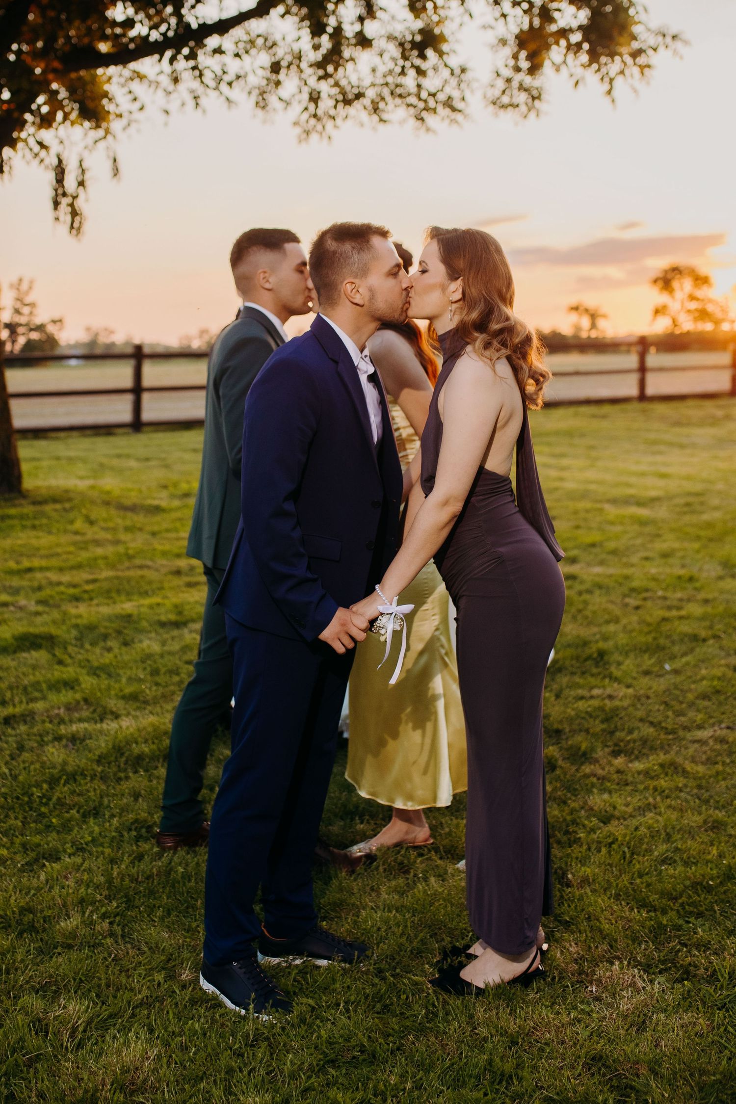 Romantic sunset photo of couple sharing a kiss on grass field with wooden fence and golden sky in background.