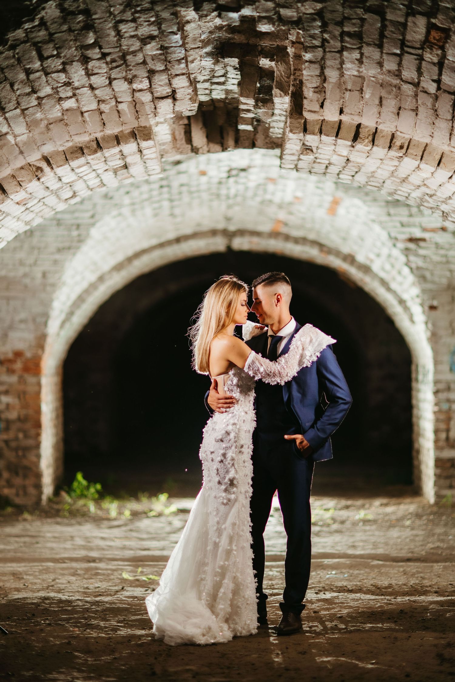 A romantic wedding portrait under an arched brick tunnel with dramatic lighting.