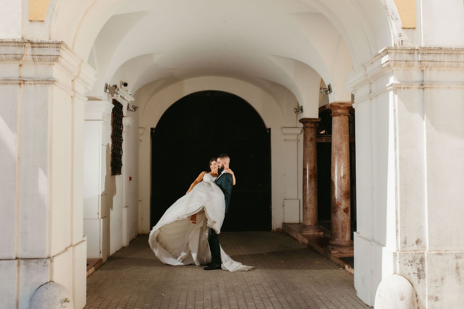 A elegant bride and groom embrace in a stunning white columned archway at a historic building venue.