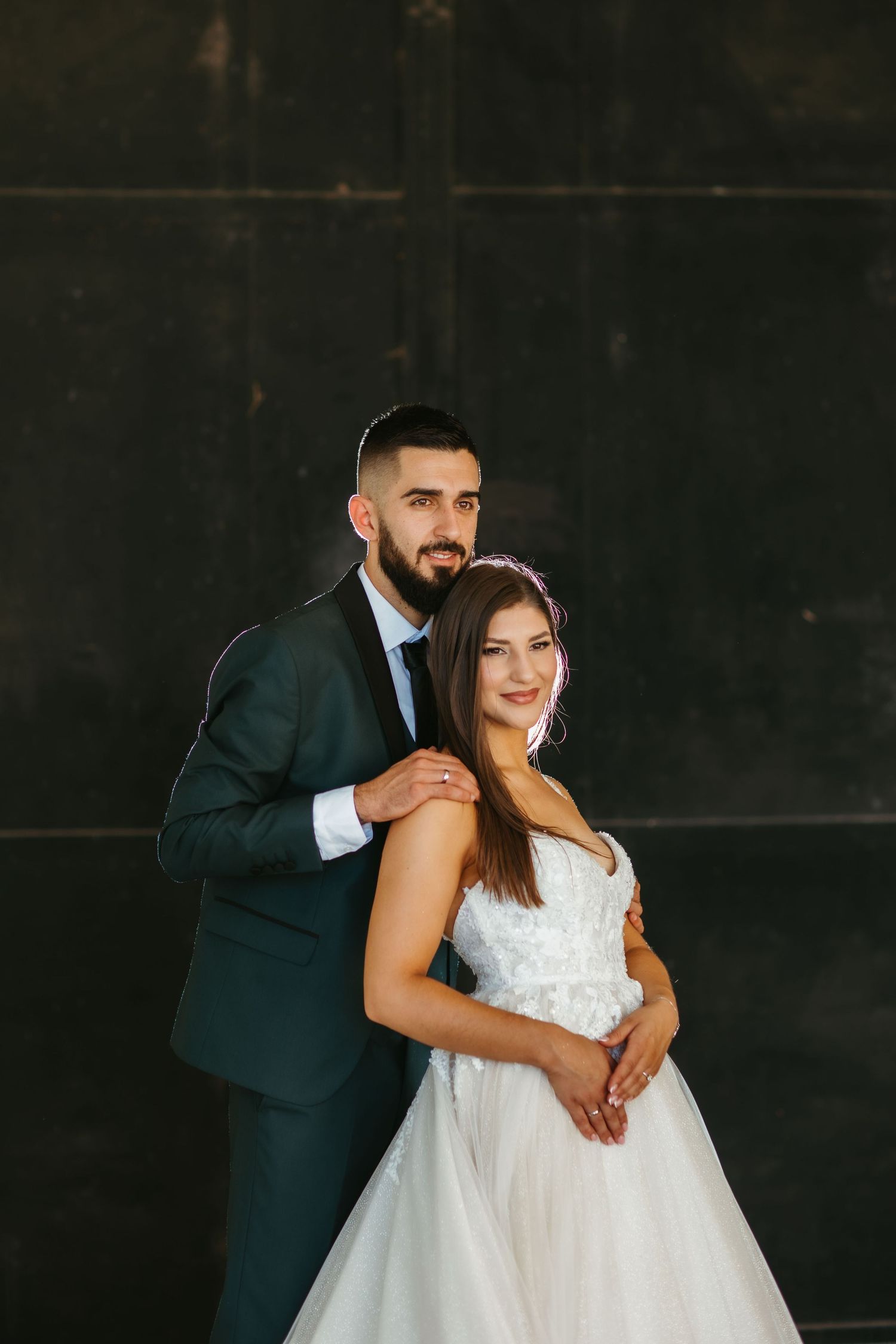 A couple poses together against a dark wall, with the bride wearing a white gown and the groom in a dark green suit.