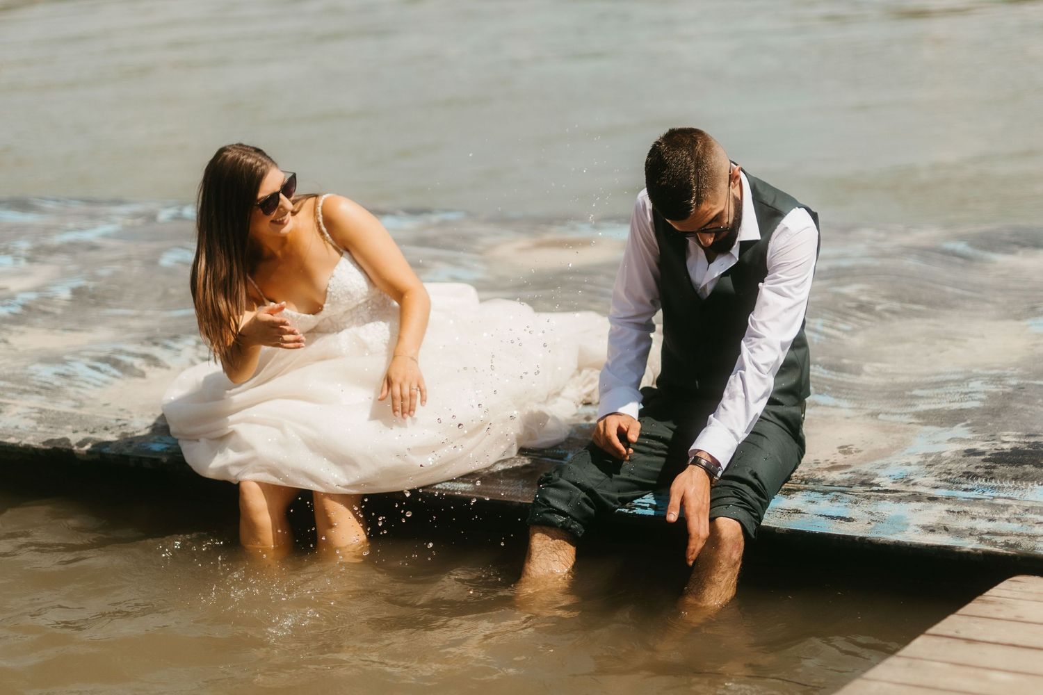 A couple in formal wedding attire sitting at the edge of water with their feet dipped in, enjoying a playful moment.