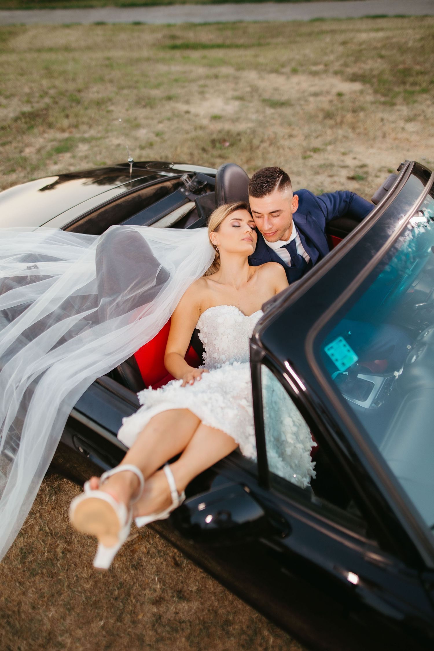 Intimate wedding portrait inside black convertible with flowing veil and formal attire.
